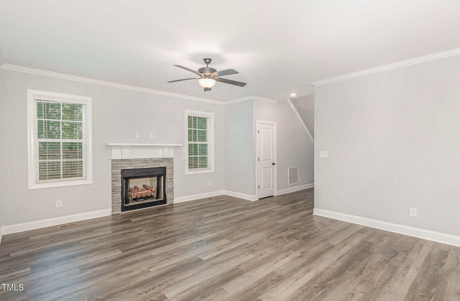 A bright, empty living room with light gray walls and wood flooring. It features a ceiling fan, a white-trimmed gas fireplace with a stone surround, two windows, and a closed door. A staircase is visible on the right.
