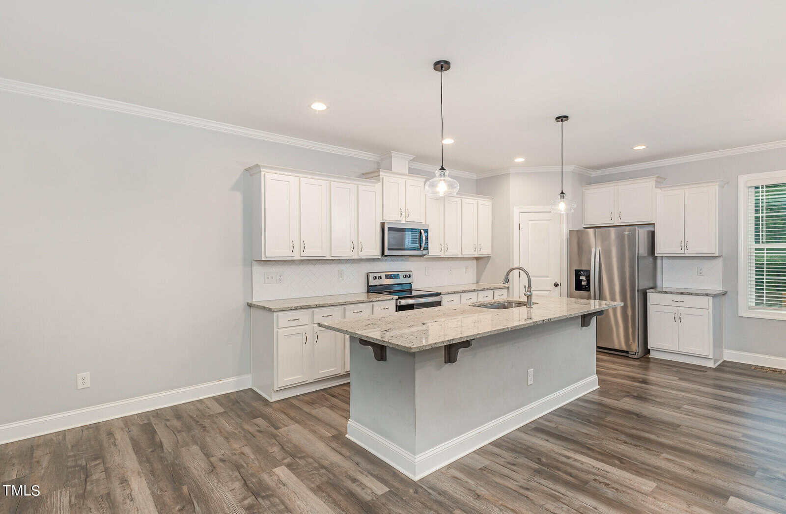 Modern kitchen with white cabinets, stainless steel appliances, and a large central island with a granite countertop. Two pendant lights hang above the island. Wooden flooring adds warmth, and a window offers natural light.
