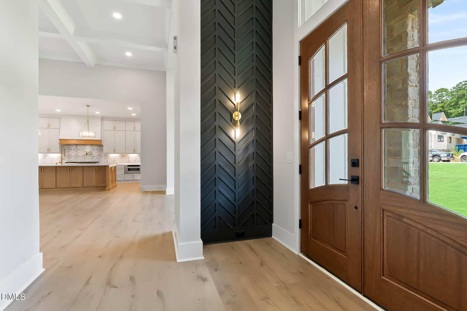 Modern home entryway with a wooden double door, black chevron accent wall with a sconce, light wood flooring, and a view into a spacious white kitchen with pendant lighting and large windows.