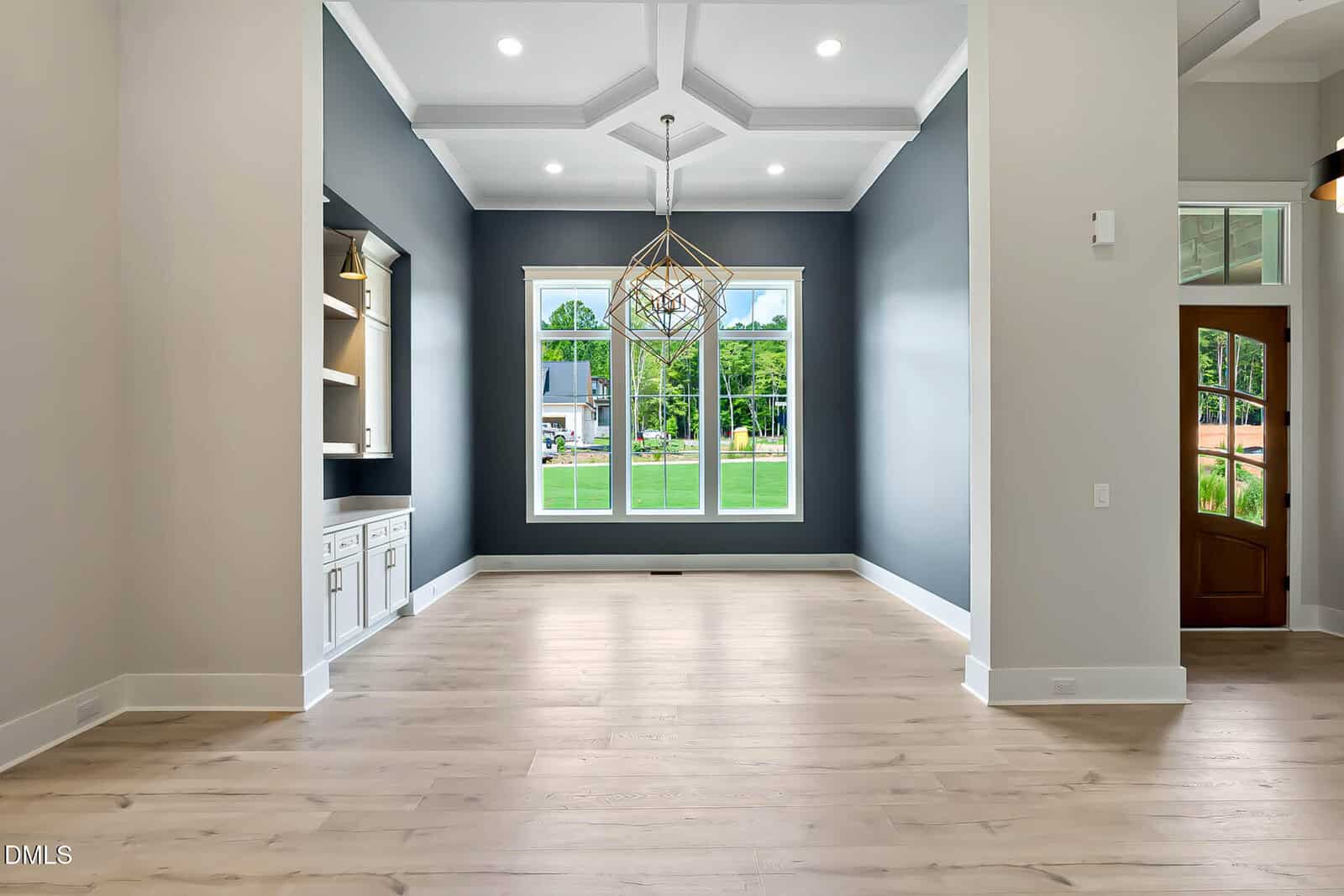 Modern dining room with light wood floors, dark blue accent walls, built-in cabinets on the left, large windows showing green lawn and houses outside, geometric chandelier, and wooden front door visible on the right.