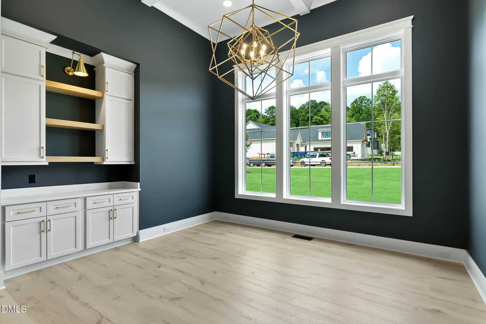 A modern empty room with light wood floors, dark gray walls, white trim, a gold geometric chandelier, large windows, built-in white cabinets, and wood shelves. Outside, a suburban neighborhood is visible.