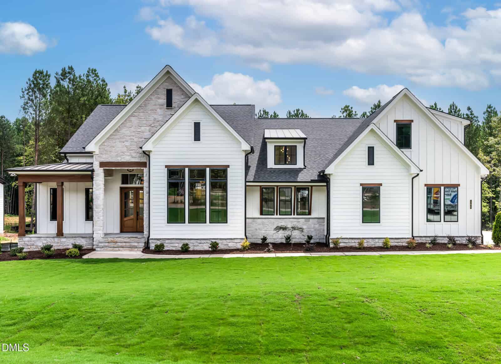 Modern single-story house with white siding, stone accents, large windows, a covered porch, and black roof at 180 Cherry Bark Drive in Youngsville. The lawn is neatly manicured, and pine trees line the background under a partly cloudy sky.