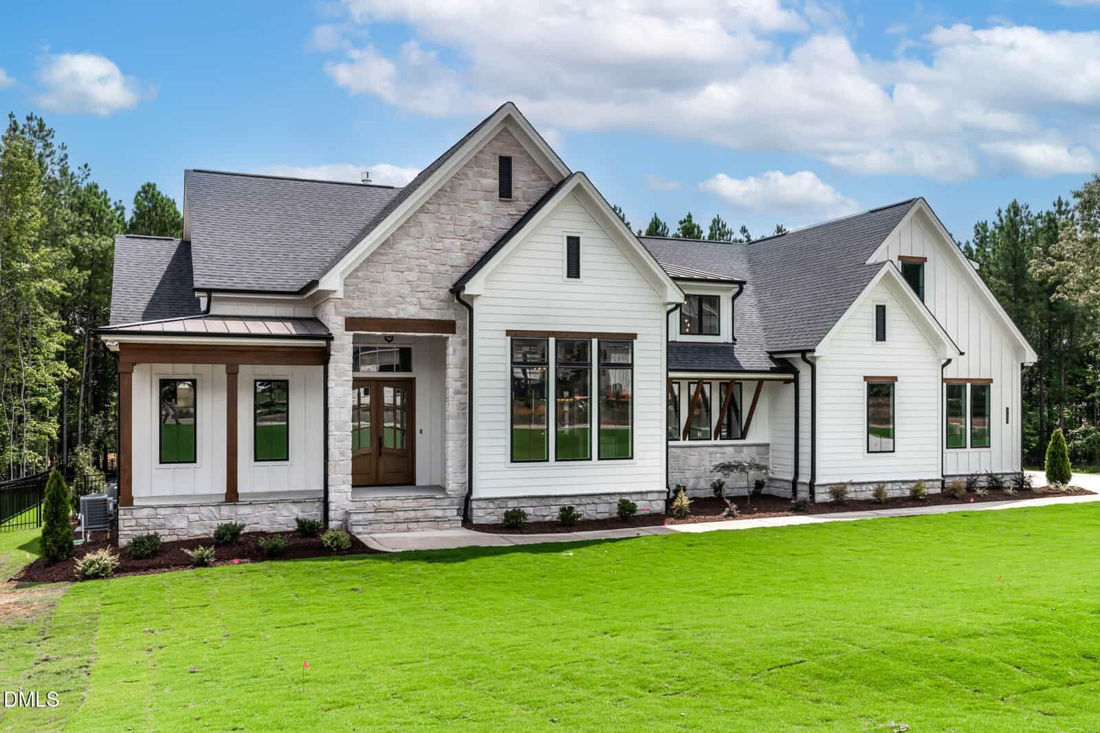 A modern two-story house with white siding, stone accents, multiple peaked roofs, large windows, and a covered entry, set against a backdrop of tall trees with a neatly manicured green lawn in the foreground.