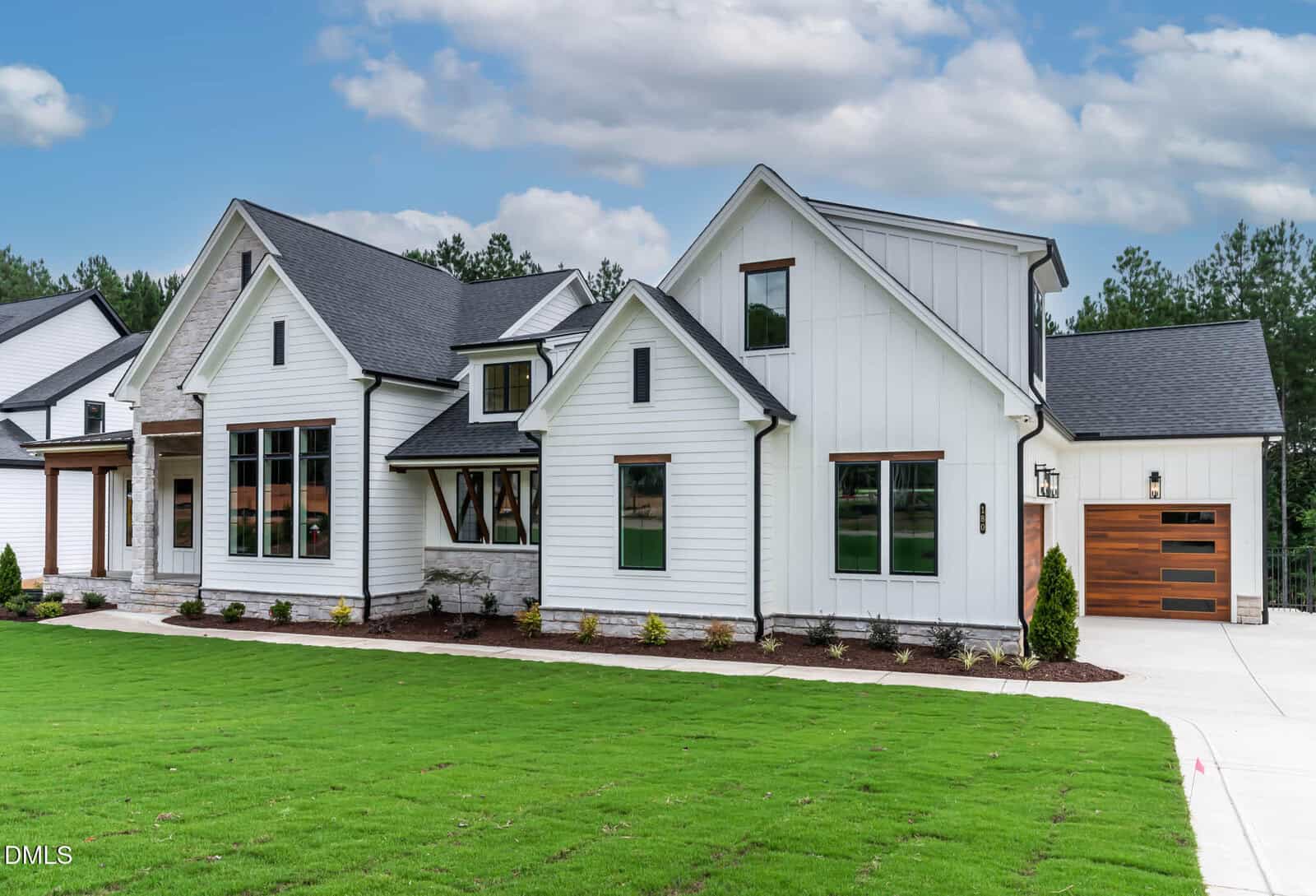 Modern white house with black roof accents, large windows, and wooden garage door sits on a well-manicured lawn with minimal landscaping, under a partly cloudy sky.