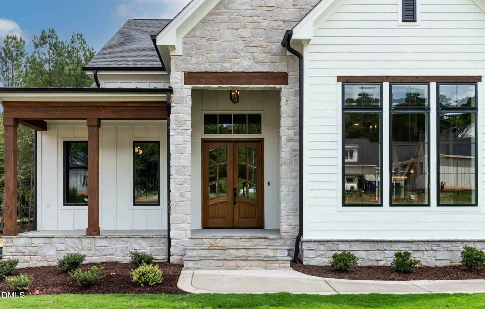 Front view of a modern house featuring light stone and white siding, wooden trim, a wooden double front door, large rectangular windows, small shrubs, and a concrete walkway. Neighboring homes are reflected in the windows.