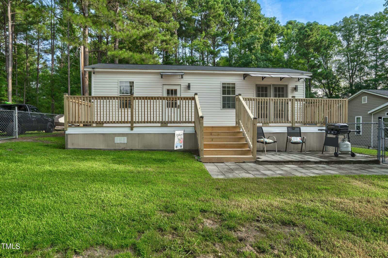 A small white house with a wooden deck and stairs, two grills, and a patio area in the backyard, surrounded by green grass and trees. A chain-link fence and a neighboring house are visible in the background.