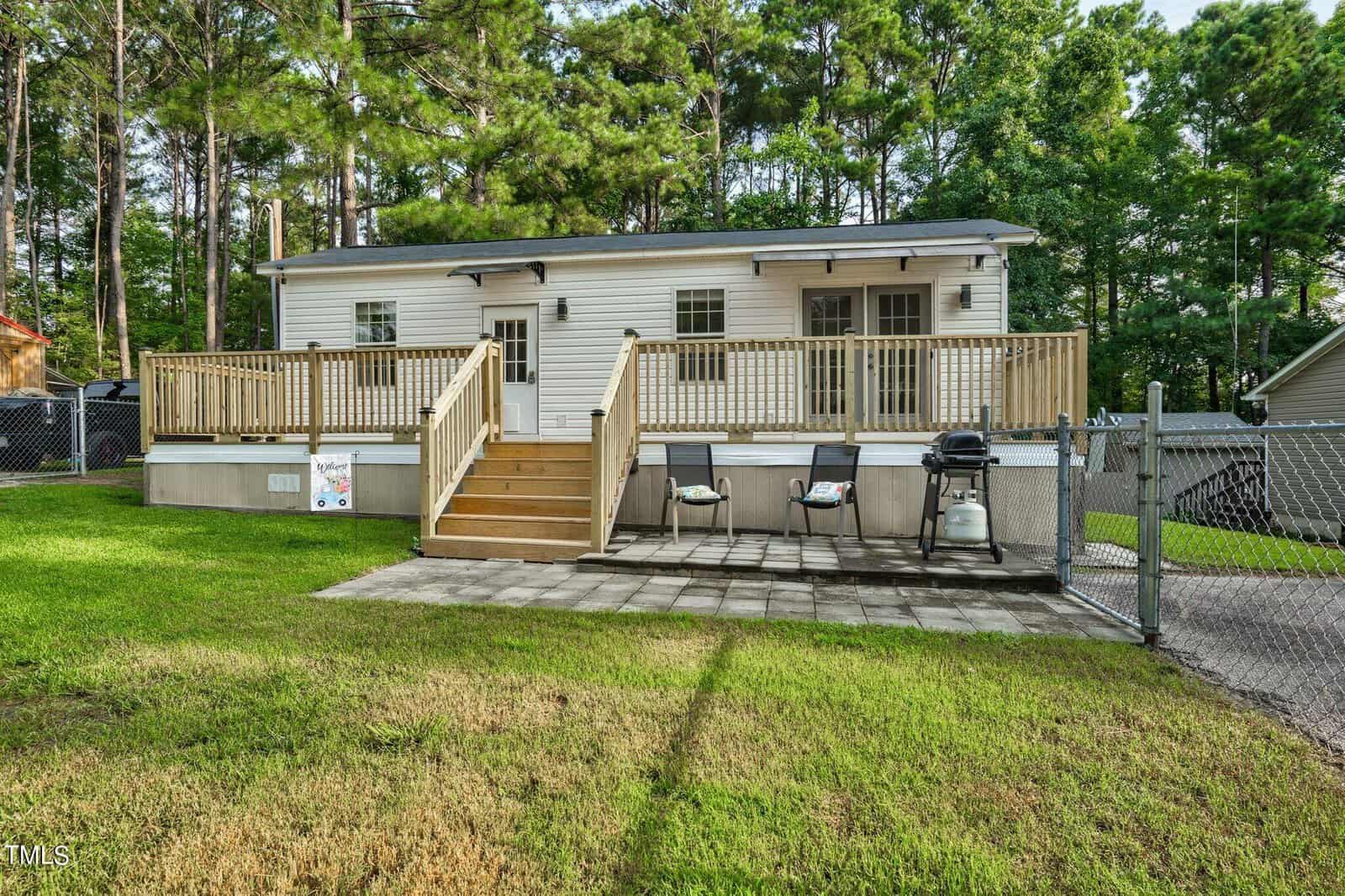 A small white house with a wooden deck, steps leading to a fenced yard, and two grills on a stone patio. The house is surrounded by green grass and tall trees in the background.