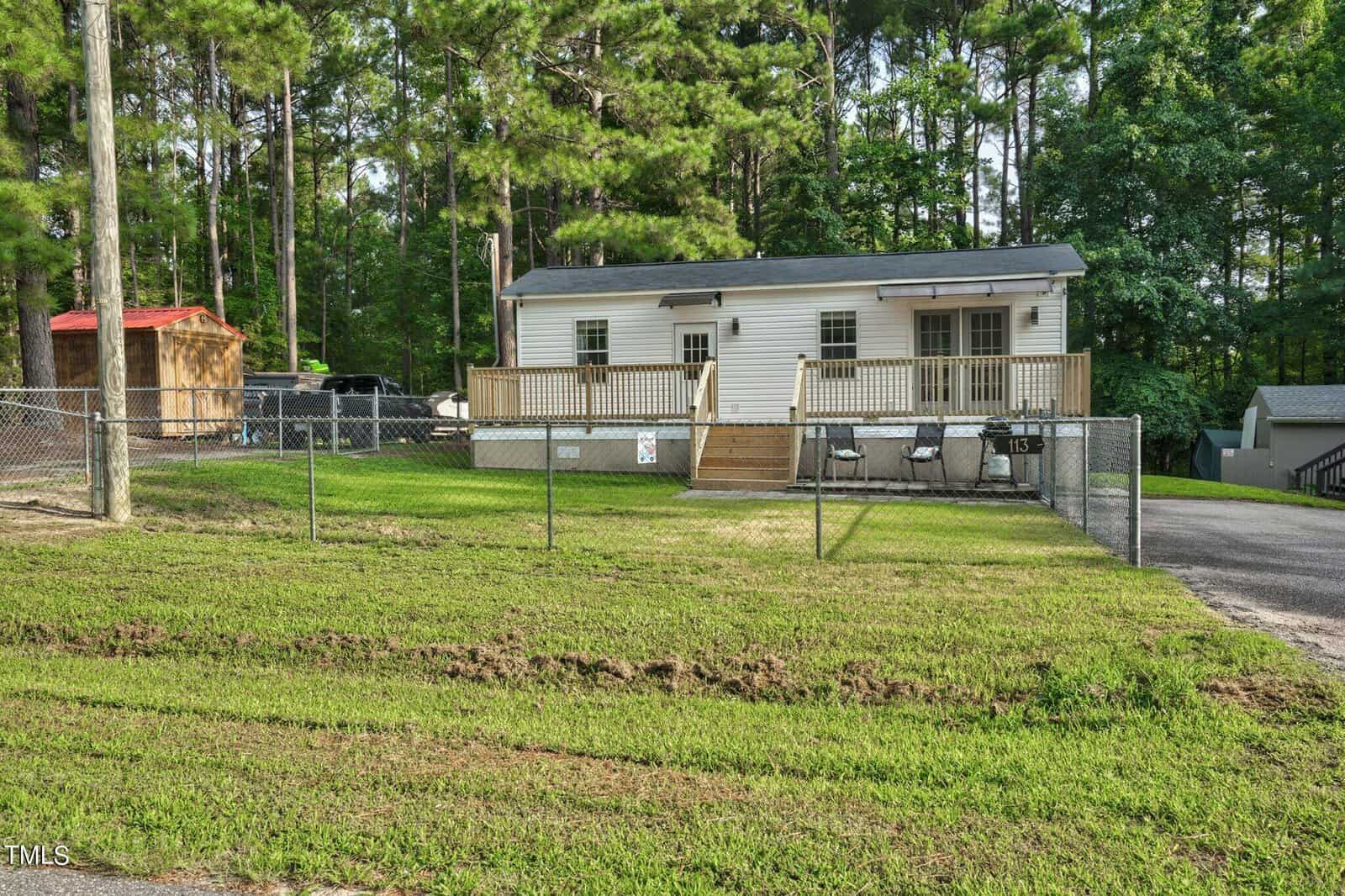 A small white house with a front porch and ramp sits behind a chain-link fence, surrounded by green grass and tall pine trees. A shed and driveway are visible to the left and right of the house.