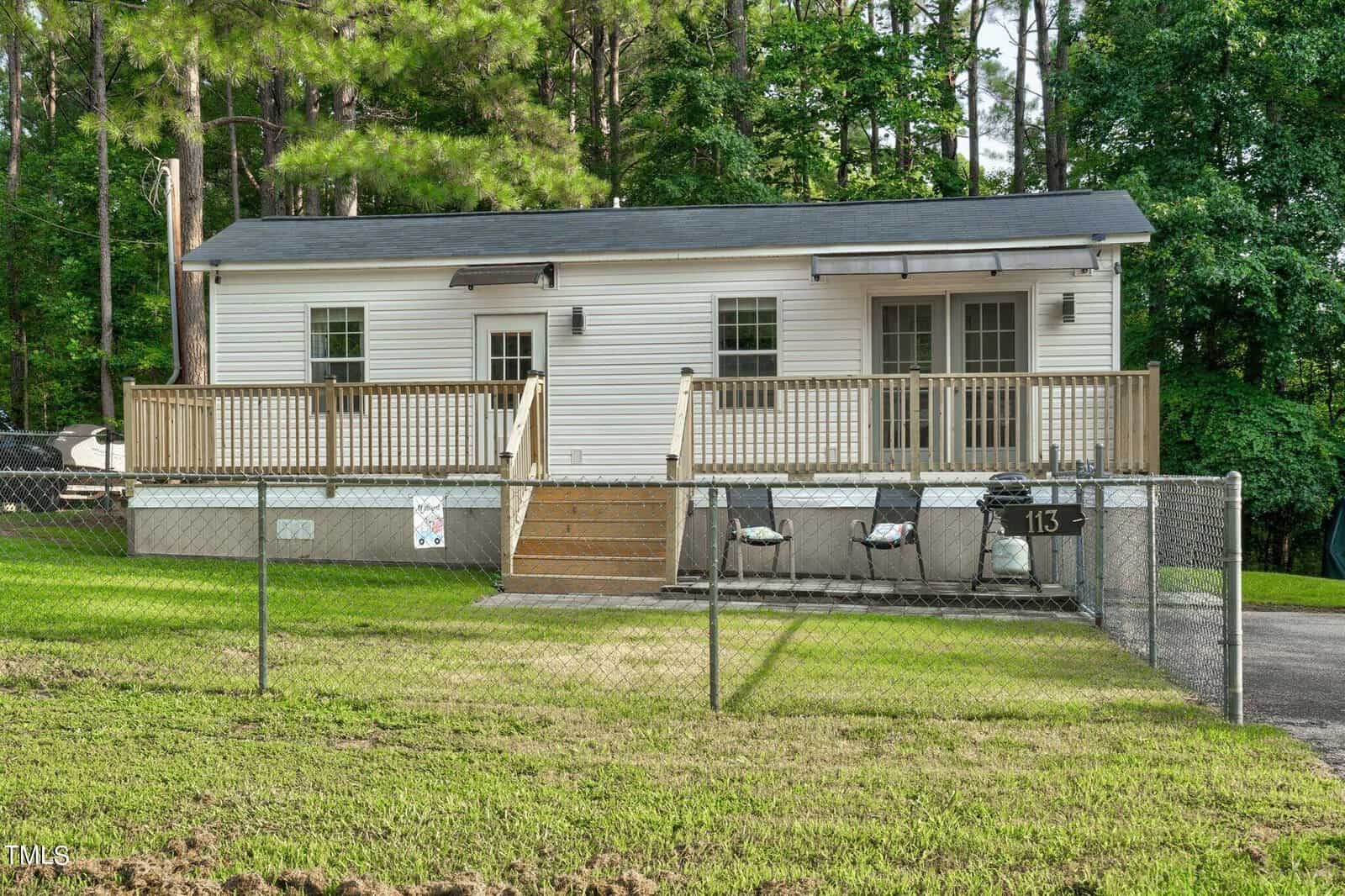 A small white house with a dark roof, wooden porch, and steps is surrounded by a chain-link fence. The house is set amid tall trees and greenery, with grills and a mailbox numbered 113 near the entrance.