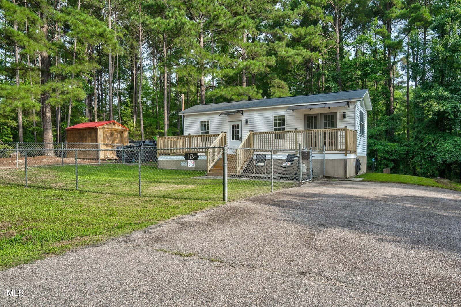 A small white house with a front porch and fenced yard sits among tall pine trees. There is a wooden shed in the yard and a paved driveway leads up to the house.