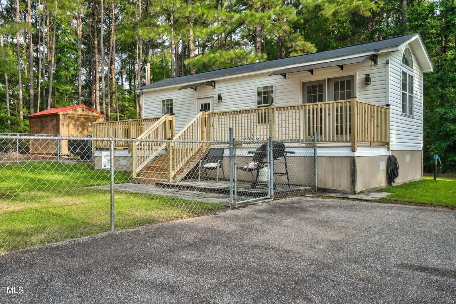 Single-story white house with a wooden porch and steps, fenced yard, outdoor seating, and a small wooden shed, surrounded by tall trees and greenery.