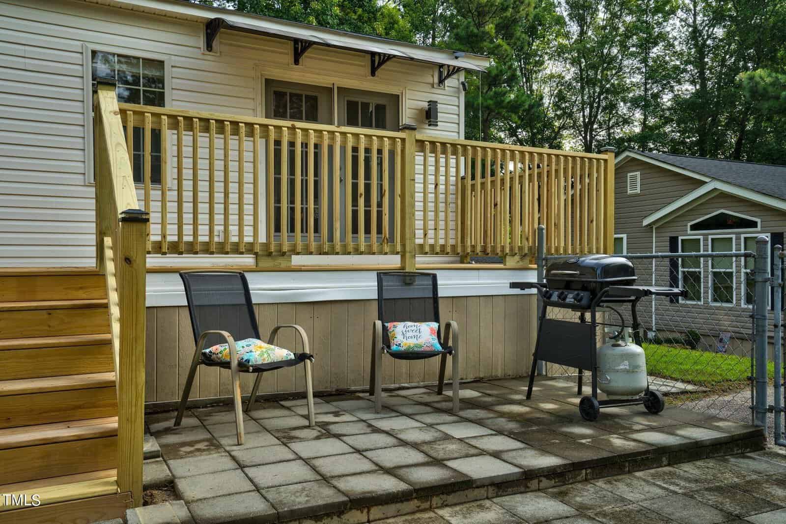 A small outdoor patio with two chairs and a propane grill on a paved area, next to wooden steps and a railing leading up to a house, with another house visible in the background.