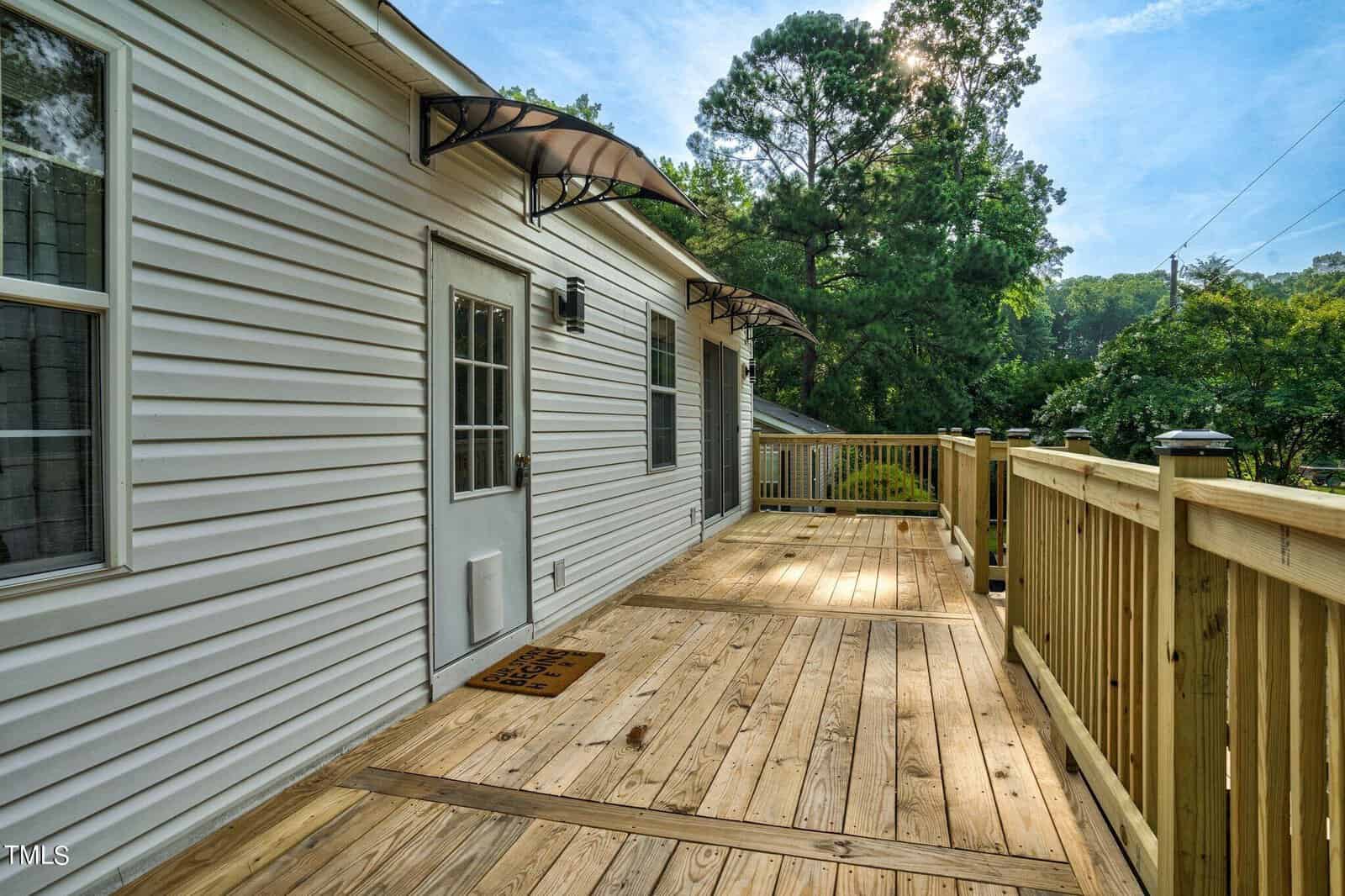 A wooden deck with a railing runs alongside a white house with several doors and awnings. Trees and greenery are visible in the background under a clear sky.