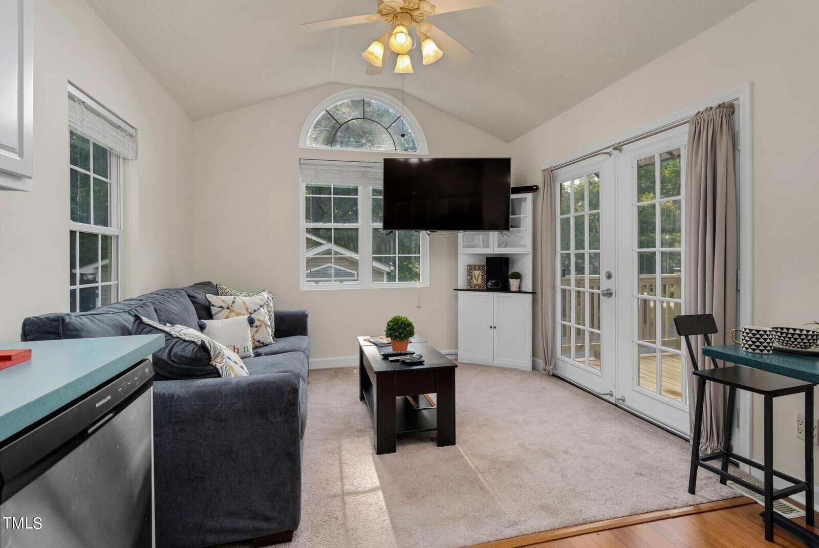 A cozy living room with a gray sectional sofa, coffee table, wall-mounted TV, and large windows and French doors letting in natural light. The space has beige carpet and light walls, with trees visible outside.