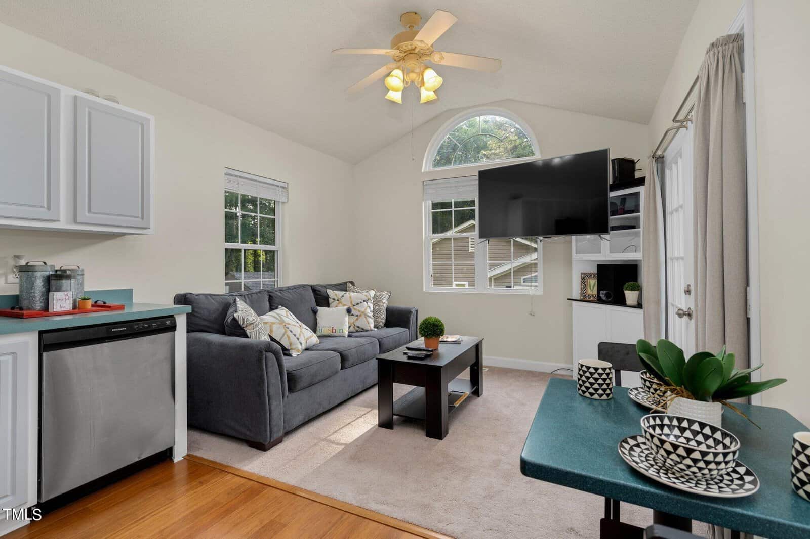Bright living room with a gray sofa, patterned pillows, coffee table, wall-mounted TV, white cabinets, and large windows bringing in natural light. A ceiling fan hangs from the vaulted ceiling. Kitchen counter with decorative bowls.