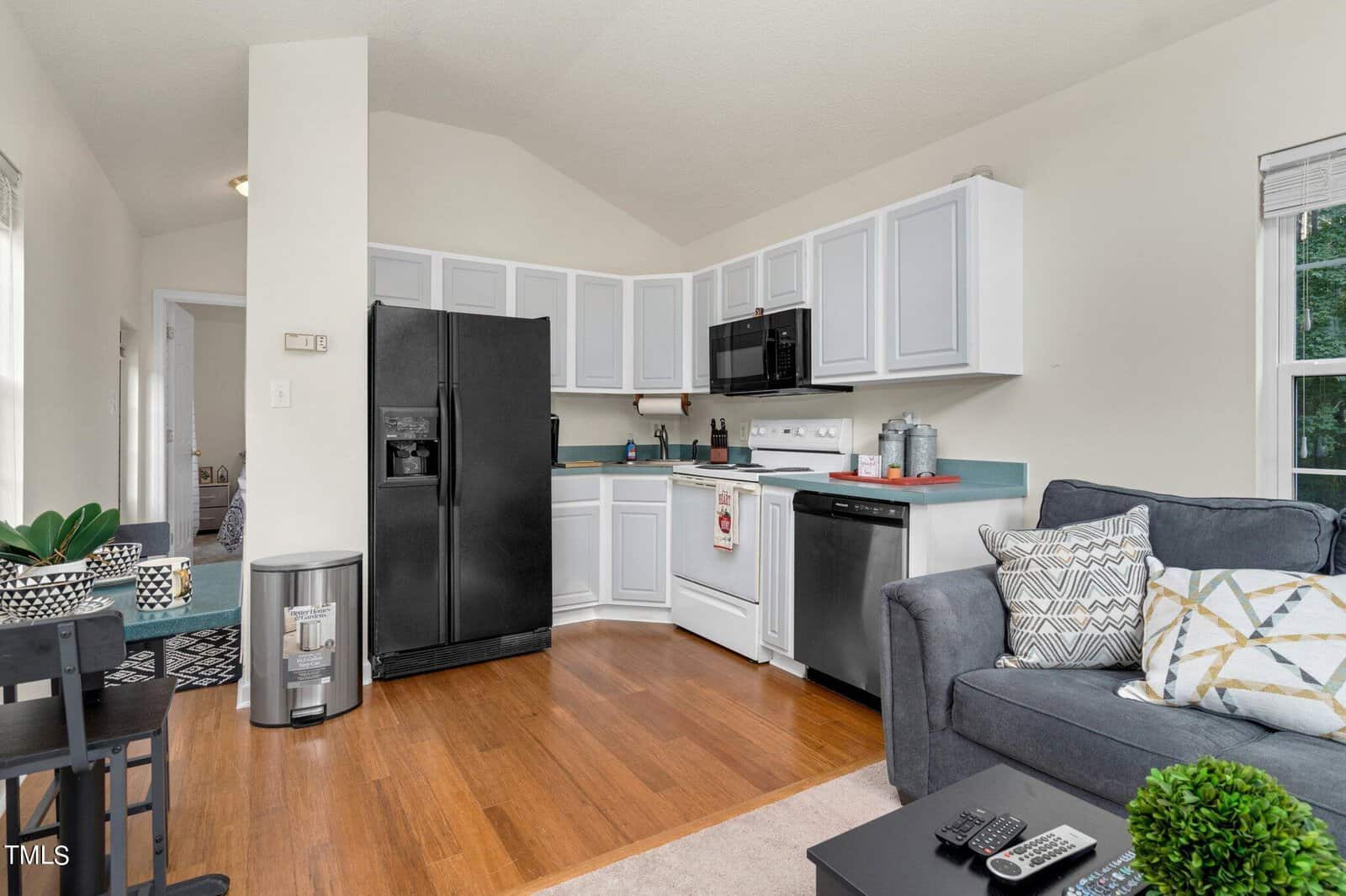 A cozy kitchen and living area with white cabinets, black appliances, a gray couch with patterned pillows, and hardwood floors. A window lets in natural light, and plants add a fresh touch.