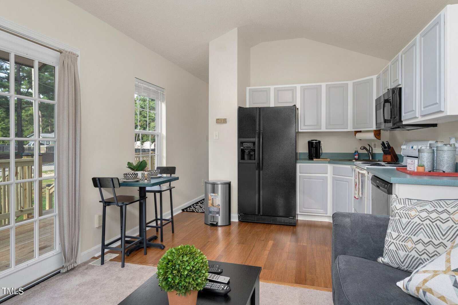Bright kitchen and dining area with white cabinets, black appliances, small dining table by the window, and a gray couch. Wood flooring and potted plants add a cozy touch.