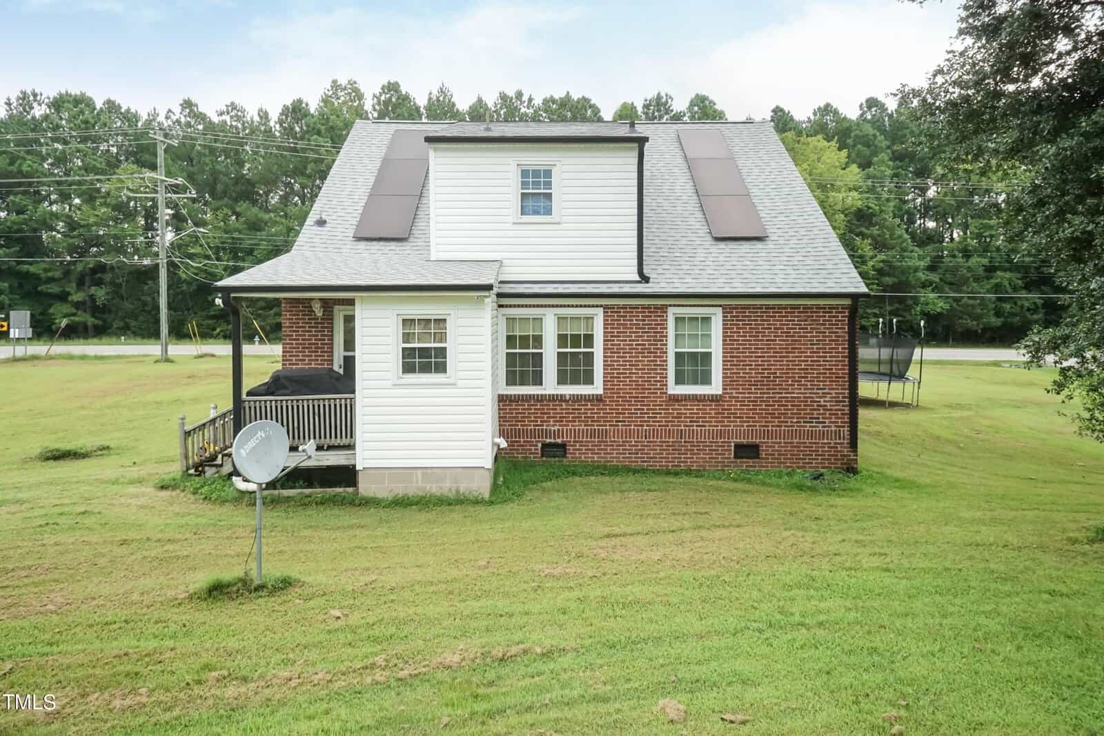 A two-story red brick and white panel house with a gray roof and solar panels. It has a satellite dish on the lawn and is surrounded by green grass and trees in the background.