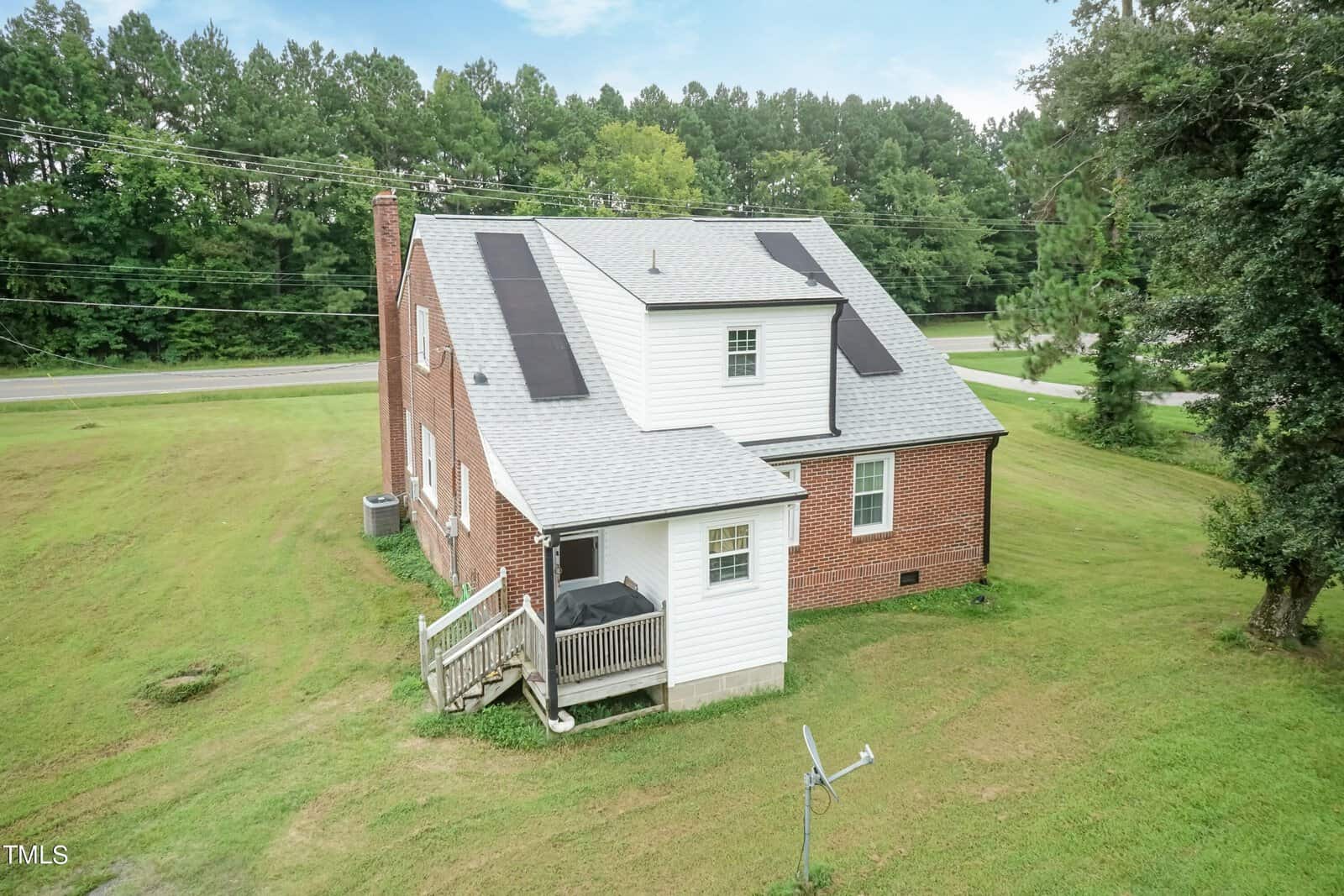 A brick house with a gray shingle roof and solar panels is situated on a spacious grassy lot. There is a small porch with stairs leading to the ground. Trees and a road are visible in the background.
