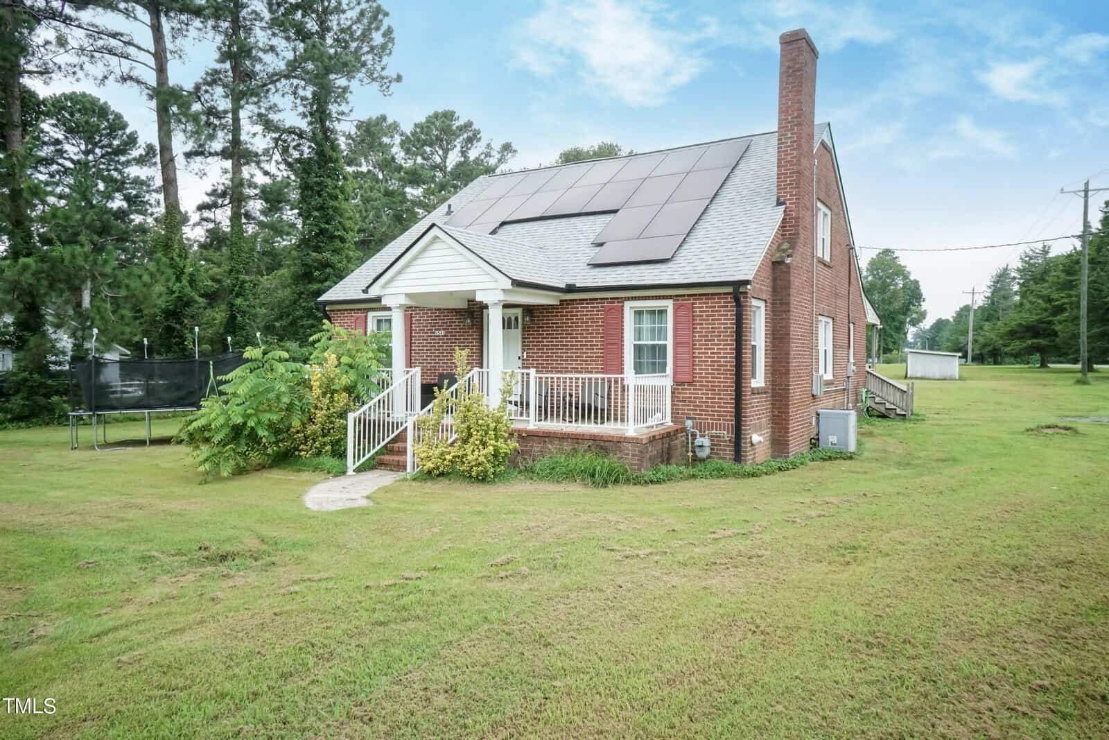 A small brick house with solar panels on the roof, a white front porch, and a chimney. Surrounded by a grassy yard with trees in the background. Overcast sky above.