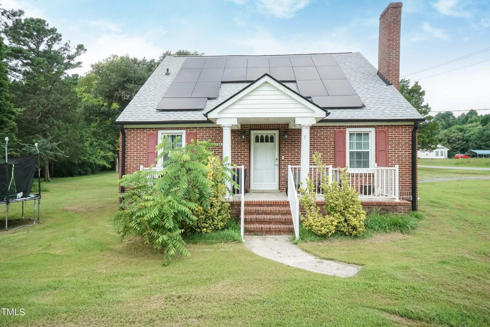 A small brick house at 1541 College Street, with a gray roof featuring solar panels, a chimney, and a white porch. A concrete path leads to the front door, surrounded by grass and shrubs. A trampoline sits left of the house. Trees and an Oxford road complete the background.
