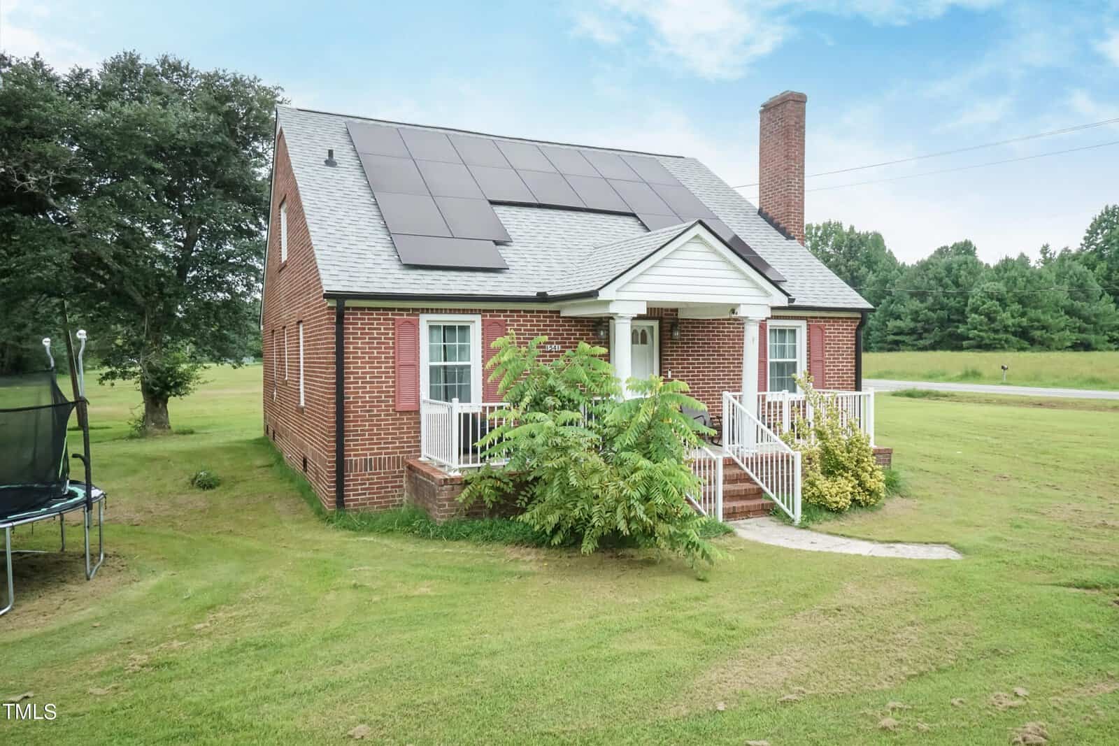 A small, brick house with solar panels on the roof, featuring a chimney and a white front porch. It's surrounded by a grassy yard with a tree to the left and a trampoline nearby. The sky is clear and blue.