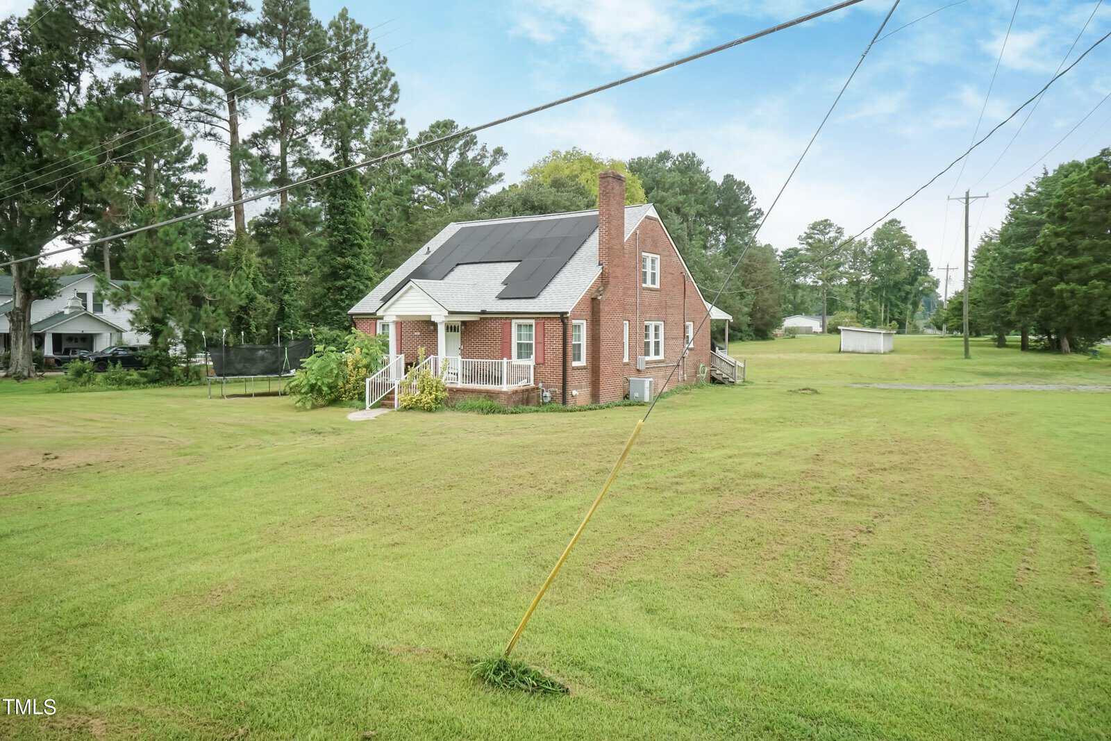 A brick house with solar panels on the roof is surrounded by a large green lawn. It features a chimney and a small front porch, with tall trees and power lines in the background under a partly cloudy sky.