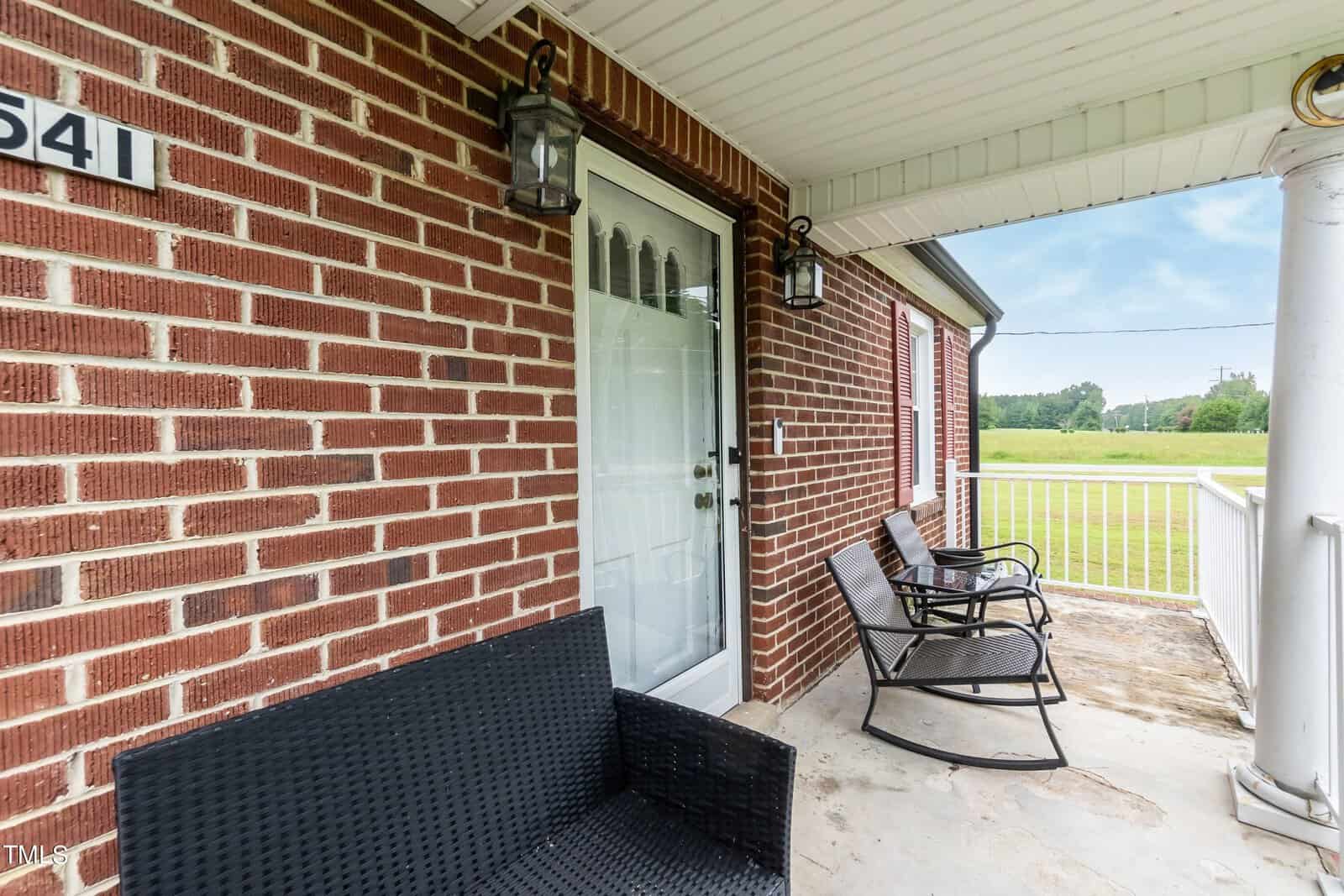 A brick porch with two black chairs and a wicker bench. A glass door is flanked by decorative lantern-style lights. The porch overlooks a grassy field, and a white column frames one side. The house number "541" is visible on the wall.