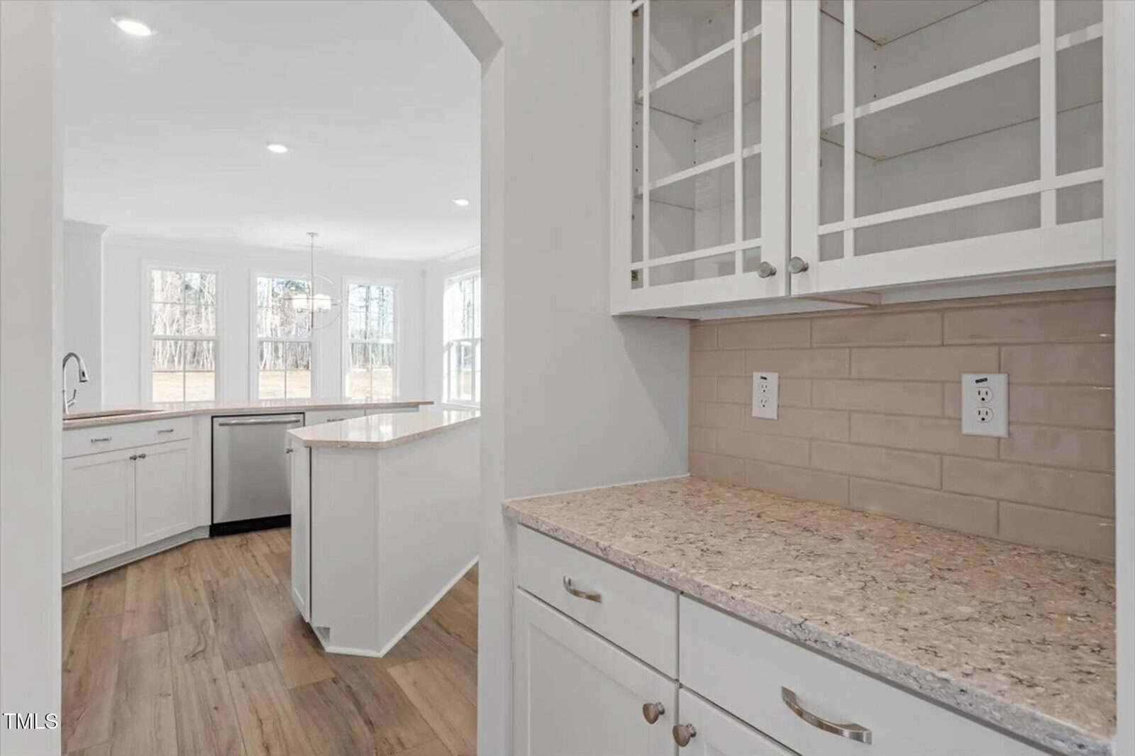 Bright modern kitchen with wood floors, white cabinets, glass-front upper cabinets, beige tiled backsplash, and granite countertops; large windows let in natural light and overlook a spacious backyard.