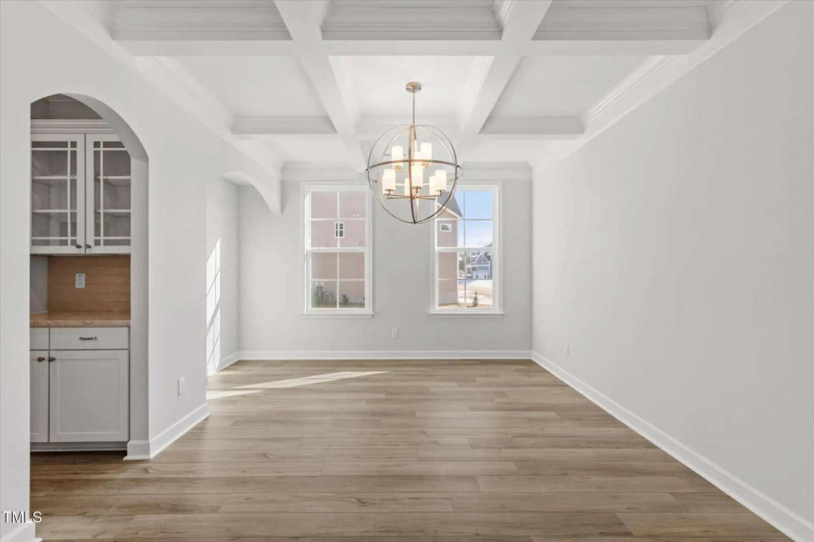 Bright, empty dining room with coffered ceiling, large windows, modern chandelier, and light wood flooring. A built-in cabinet with glass doors is visible through an arched doorway on the left.
