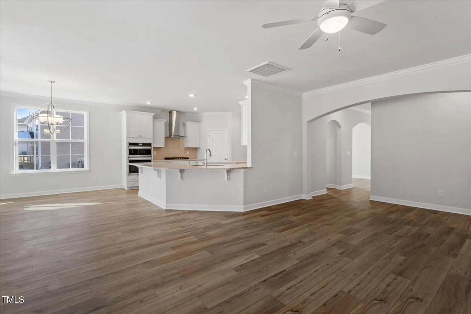 Open floor plan featuring a spacious kitchen with an island and stainless steel appliances, adjacent dining area with large window, light wood flooring, ceiling fan, and arched hallway entry.