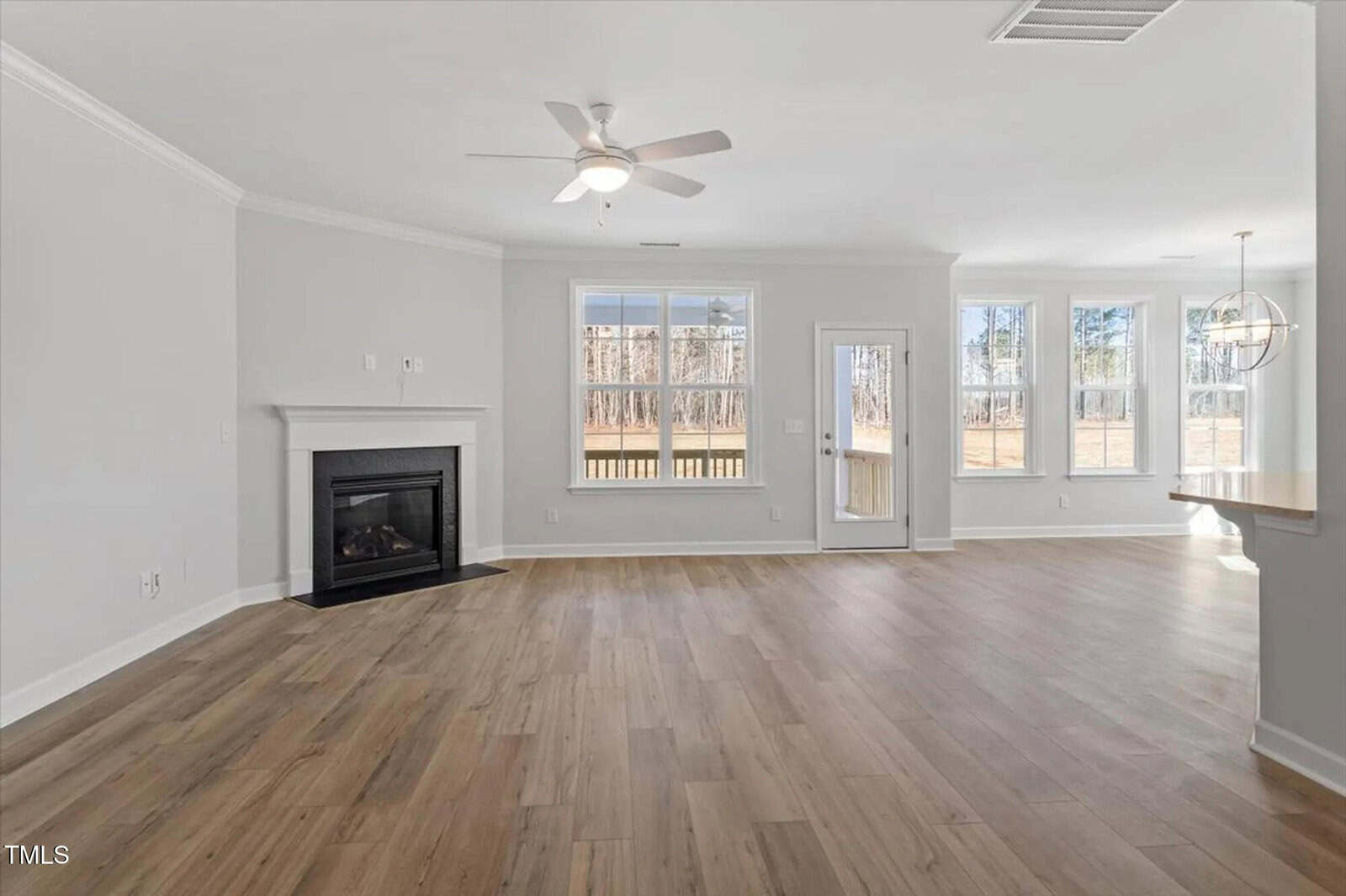 Spacious, bright living area with light wood flooring, white walls, a ceiling fan, fireplace on the left, large windows, a glass door to outside, and an open dining area with modern light fixture on the right.