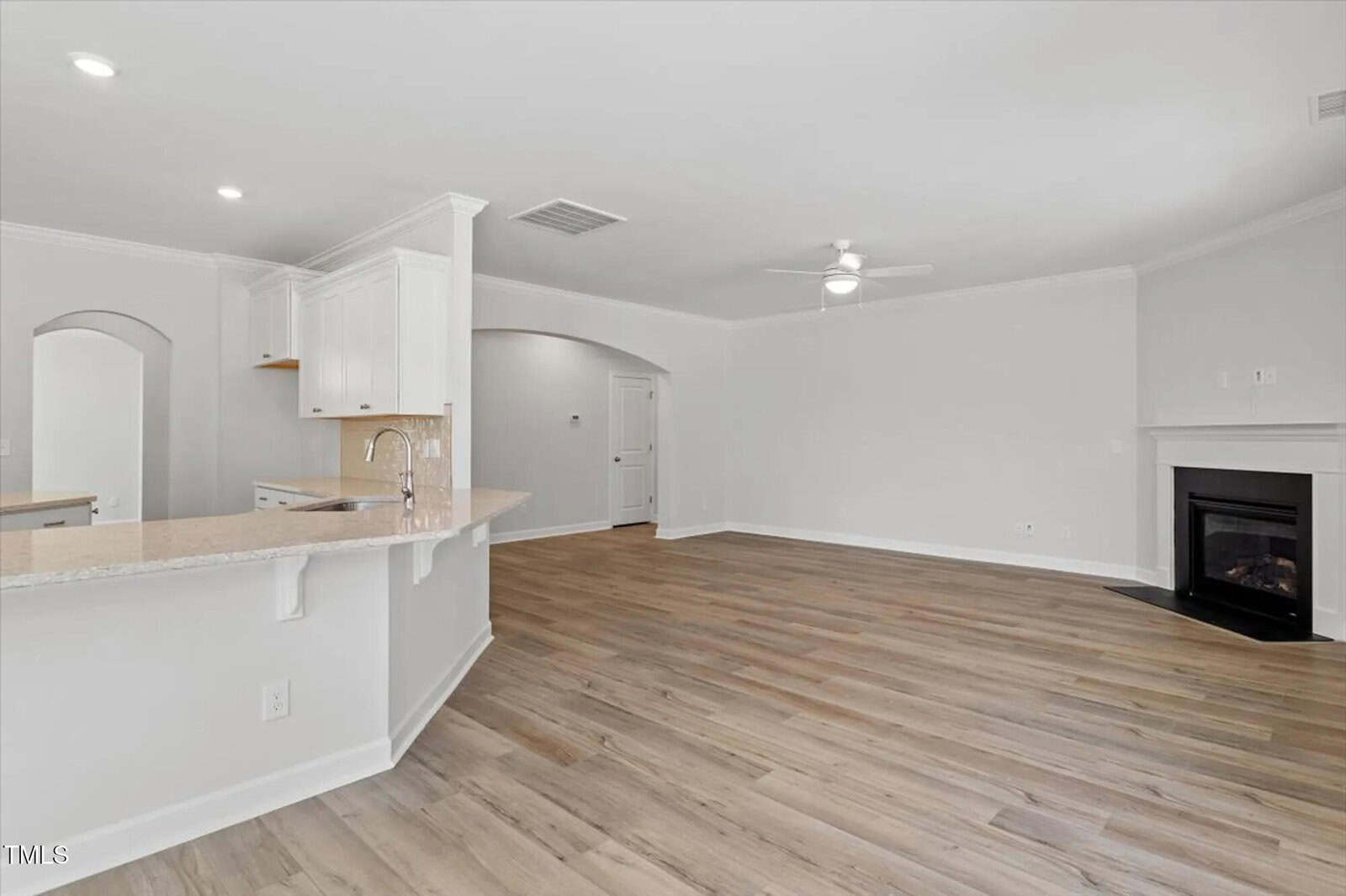 Spacious, unfurnished open-concept living area with light wood flooring, white walls, a ceiling fan, a corner fireplace, and a kitchen with white cabinets and a breakfast bar.