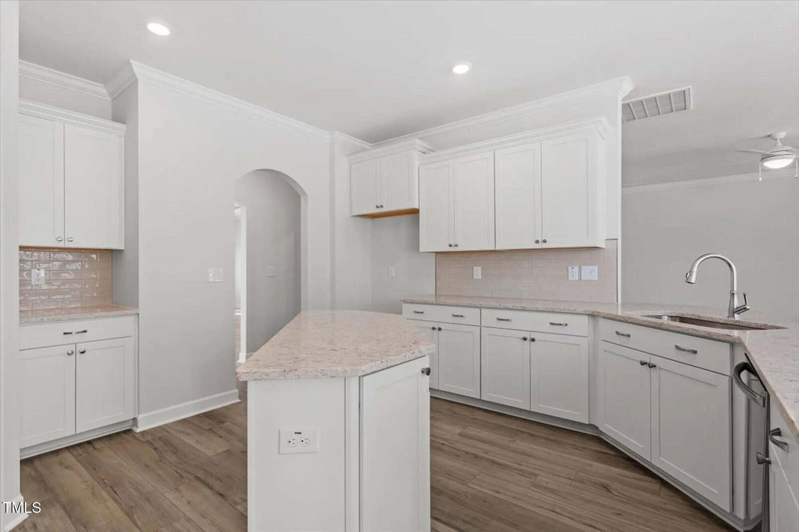 Modern kitchen with white cabinets, light-colored countertops, stainless steel sink, tile backsplash, and wood flooring. Recessed lighting and an arched doorway are also visible. The space looks clean and bright.