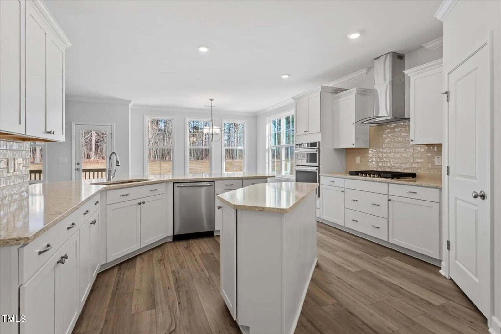Bright, modern kitchen with white cabinets, stainless steel appliances, a center island, light stone countertops, wood-look floors, and large windows offering a view of trees and a deck outside.