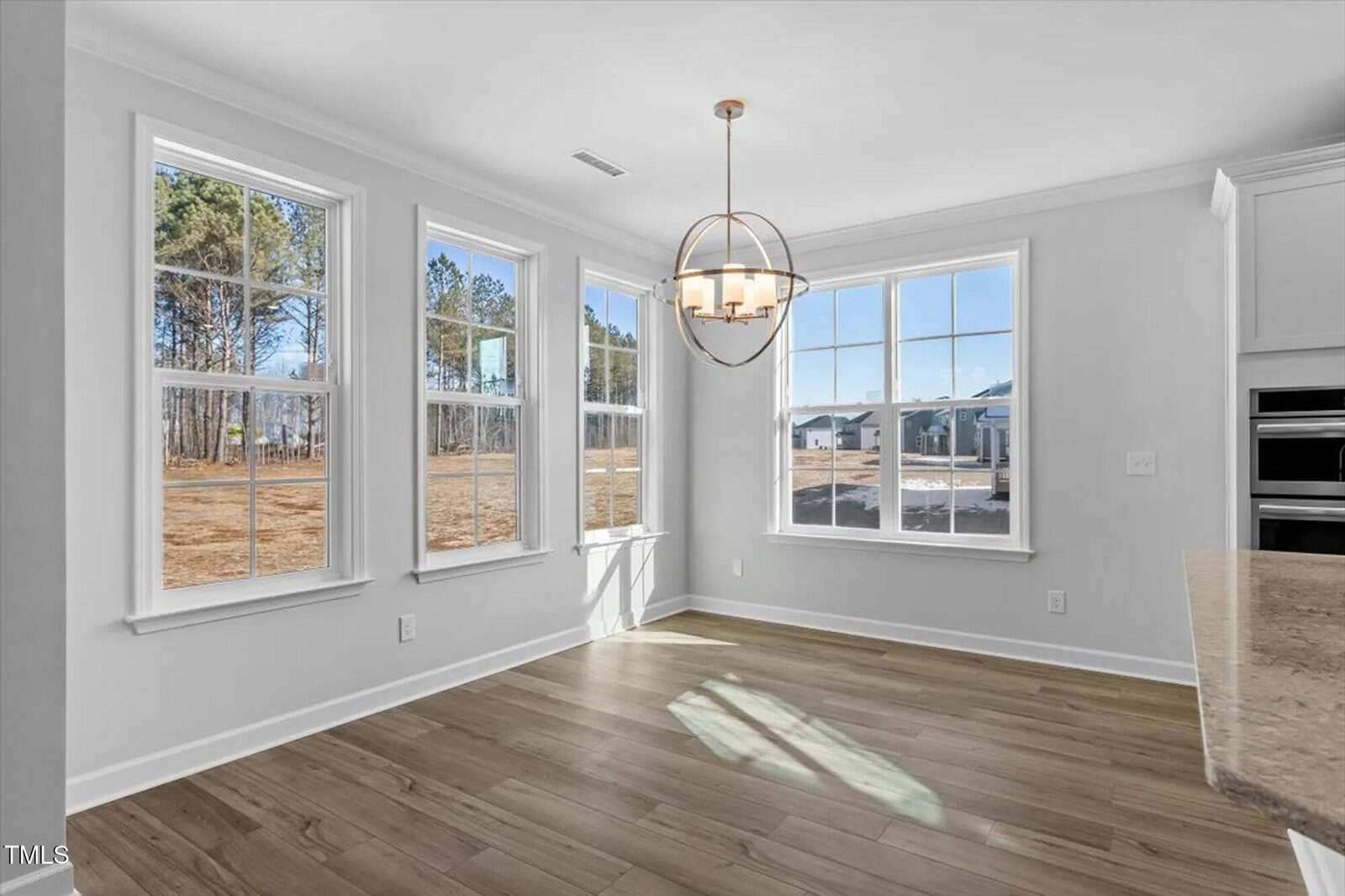 Bright dining area with large windows, light gray walls, wood-style flooring, and a modern chandelier. Sunlight pours in, showing an outdoor view of trees and a neighborhood. Part of a kitchen counter is visible on the right.
