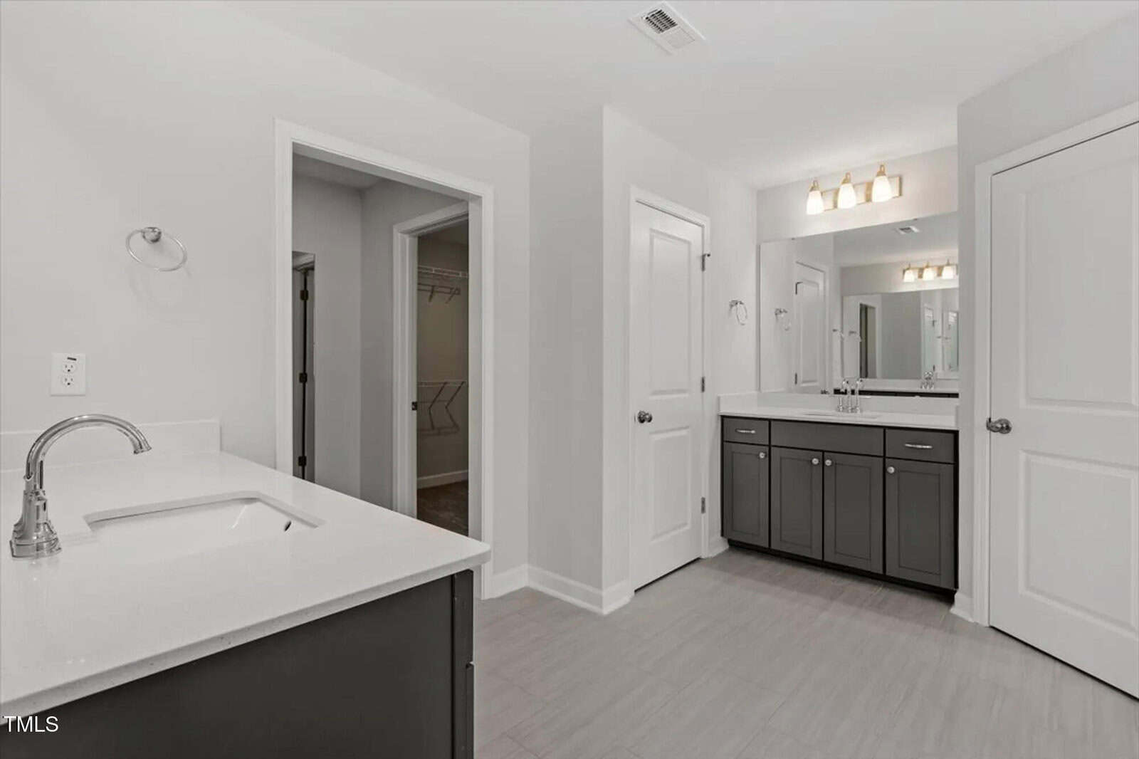 Modern bathroom with two sinks, dark cabinets, white countertops, light gray floors, and a large mirror. There are two closed doors and an open doorway leading to a walk-in closet with shelves and hanging rods.