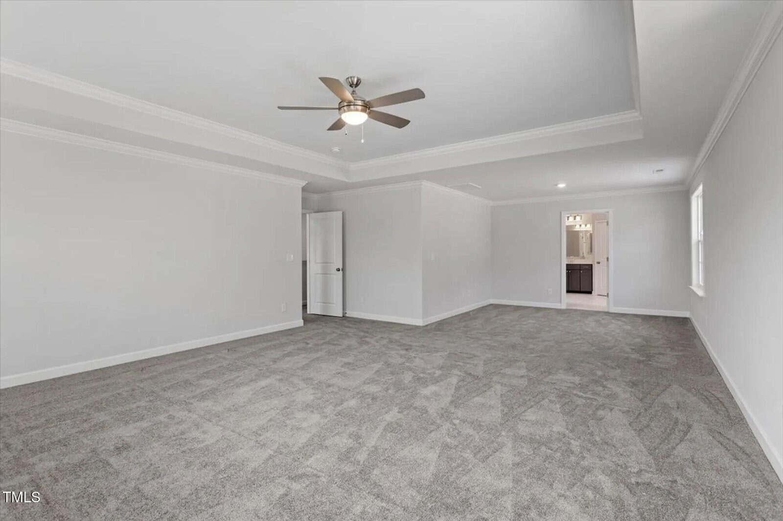 A spacious, empty room with light gray carpet, white walls, a ceiling fan with lights, and crown molding. An open doorway leads to another room with dark cabinets and a lit vanity mirror.