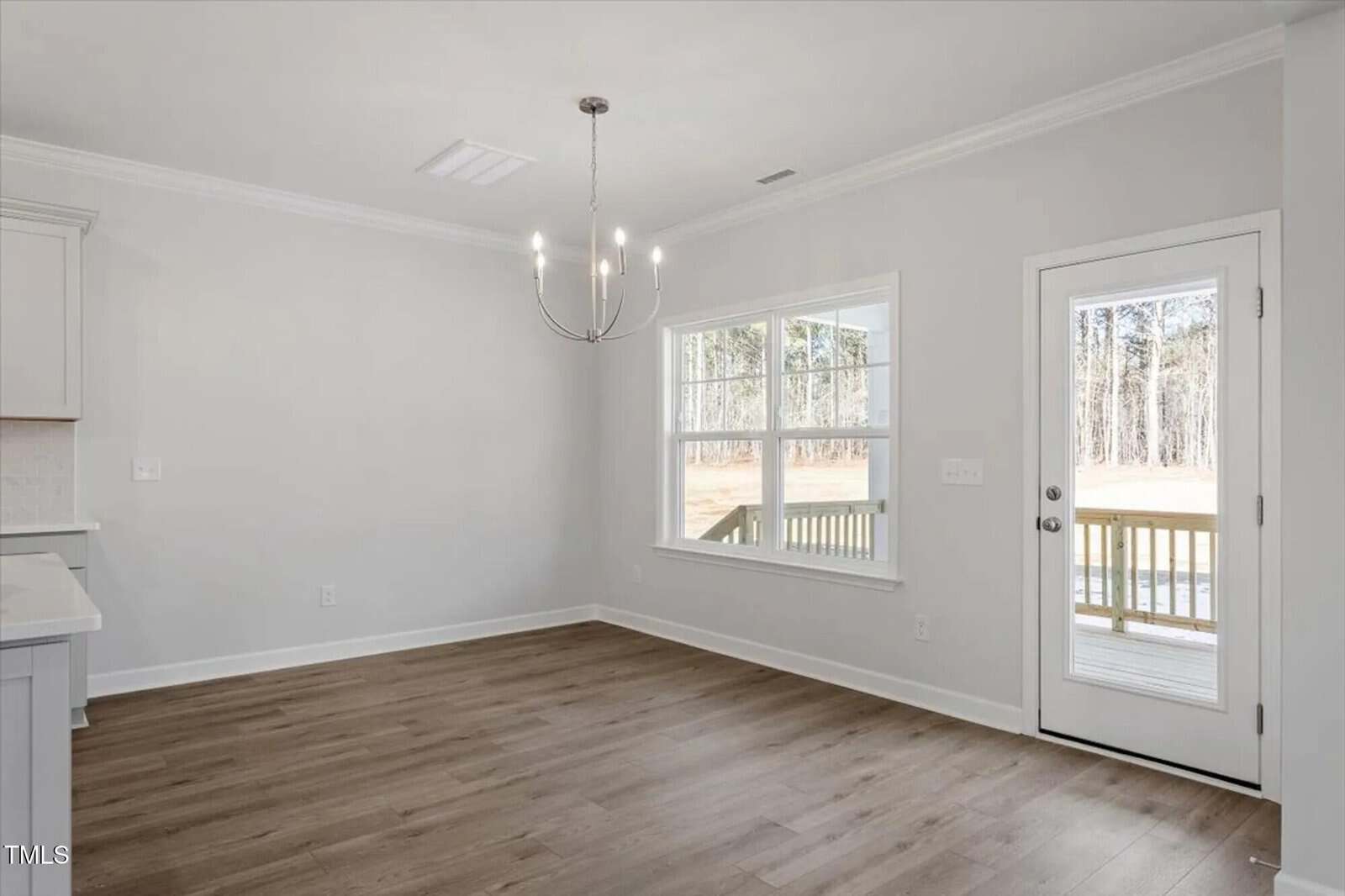 A bright, empty room with light gray walls, wood flooring, a modern chandelier, a large window, and a glass door leading to a porch with a view of trees outside.