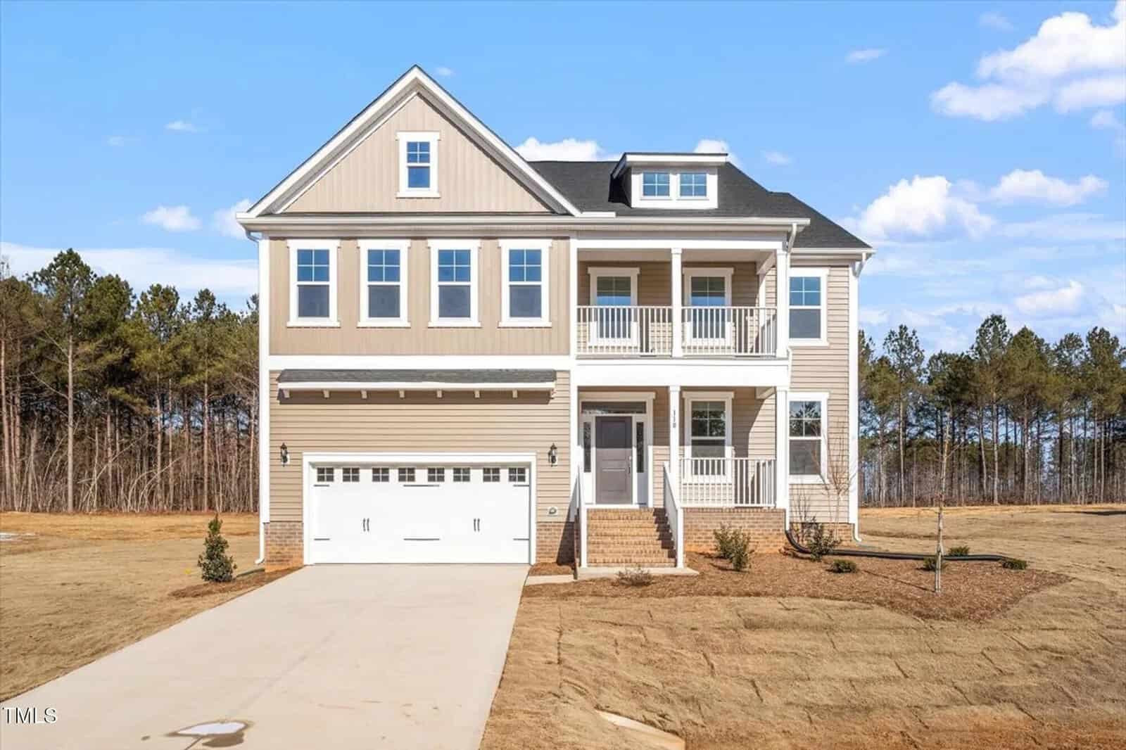 A two-story beige house with white trim, a double garage, and a covered front porch. The house sits on a newly landscaped lawn with young trees and is set against a backdrop of trees under a blue sky.
