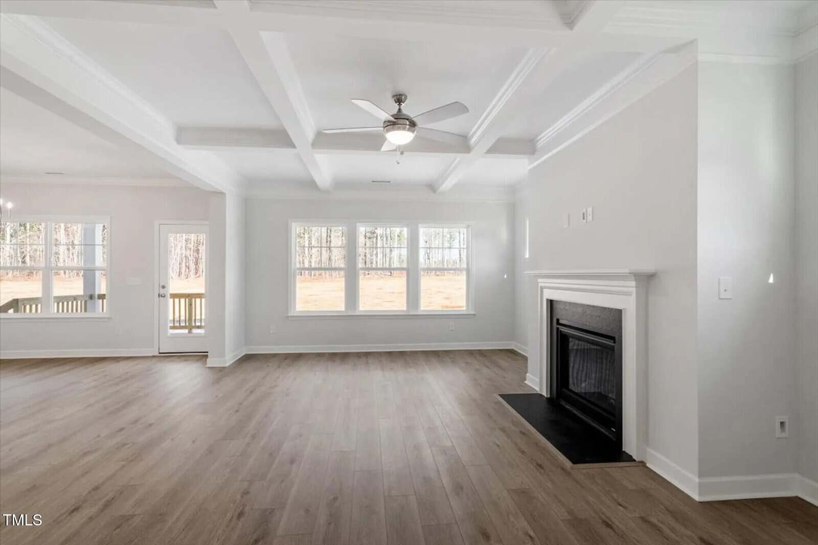 A bright, empty living room with light wood floors, large windows, a white fireplace, ceiling fan, and coffered ceiling. An open doorway leads to another room with similar flooring and natural light.