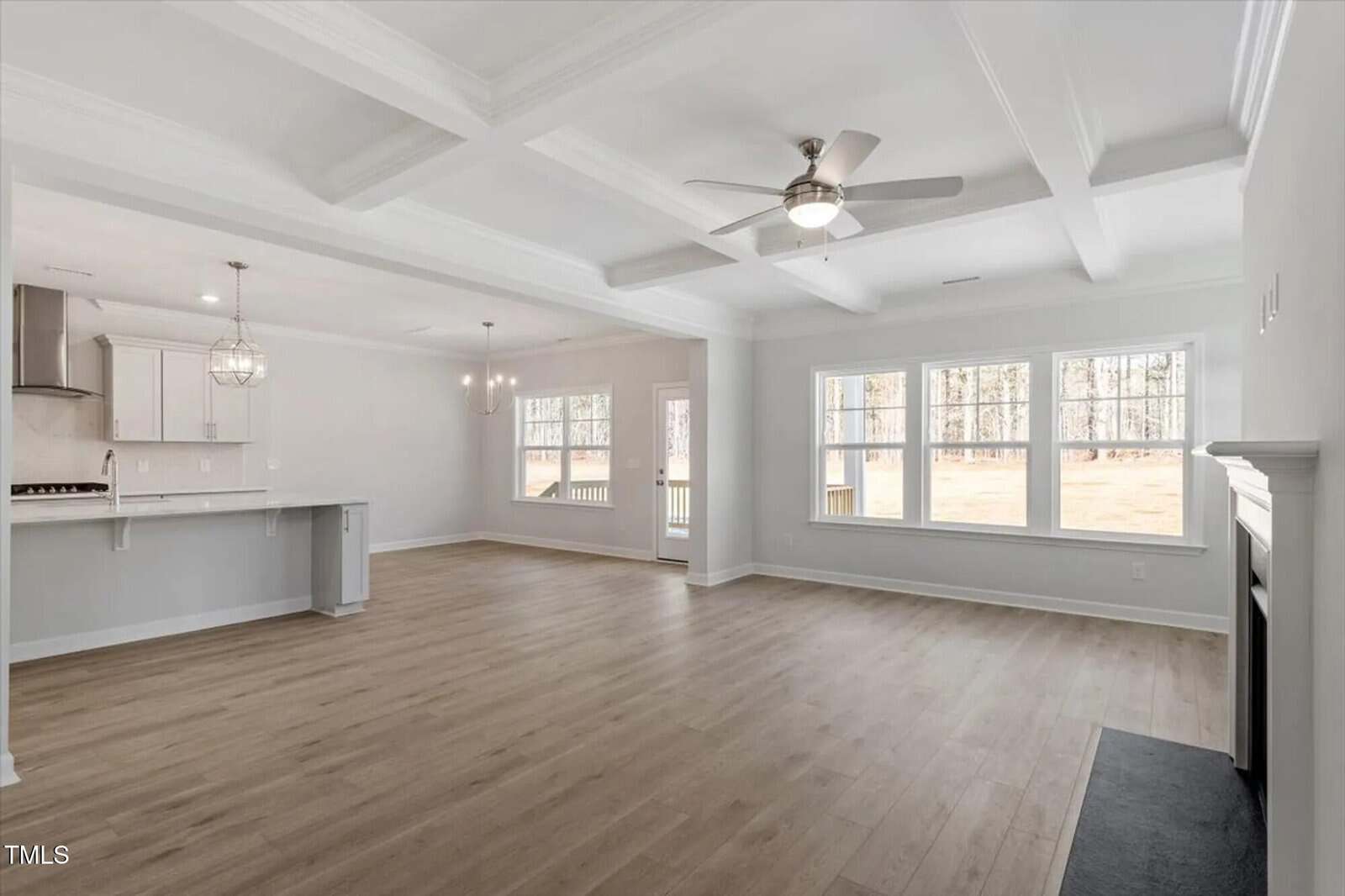 Spacious open-concept living area with light wood floors, white coffered ceiling, ceiling fan, large windows, a fireplace, and an adjacent modern kitchen with pendant lights and a breakfast bar.