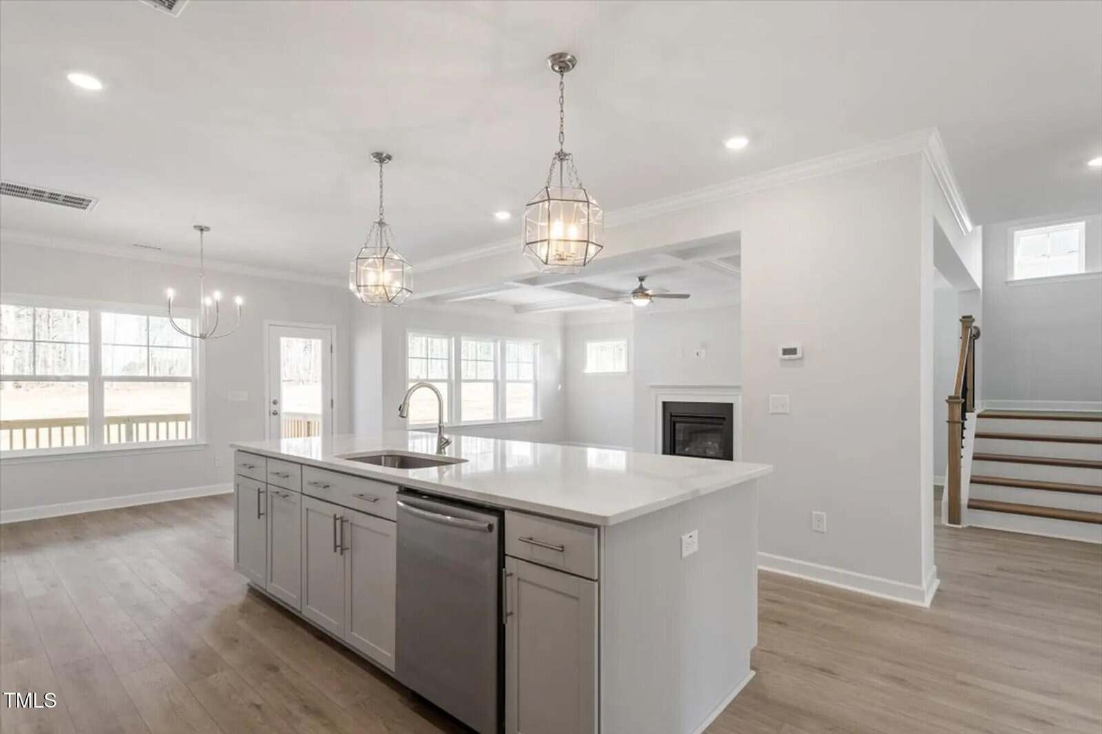 Modern, open-concept kitchen and living area with white cabinetry, stainless steel appliances, a large island, pendant lights, light wood floors, and a staircase leading upstairs. Large windows let in natural light.