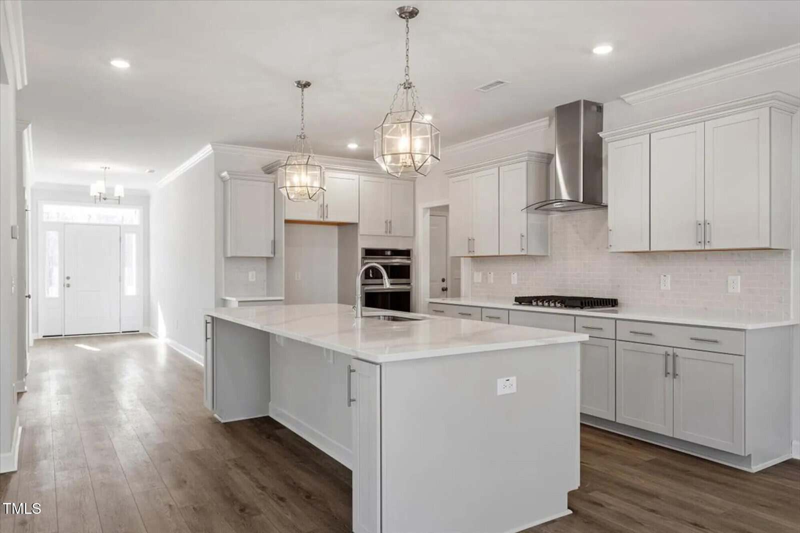 Bright, modern kitchen with white cabinets, a large island, stainless steel appliances, pendant lighting, light wood flooring, and white subway tile backsplash. An entry hallway is visible in the background.