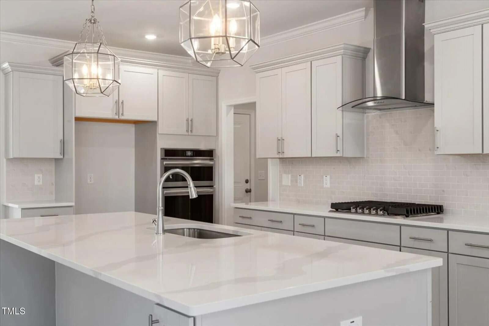 Modern kitchen with white cabinets, a large white island with a sink, stainless steel appliances, a gas cooktop with a vent hood, light gray subway tile backsplash, and two geometric pendant lights.