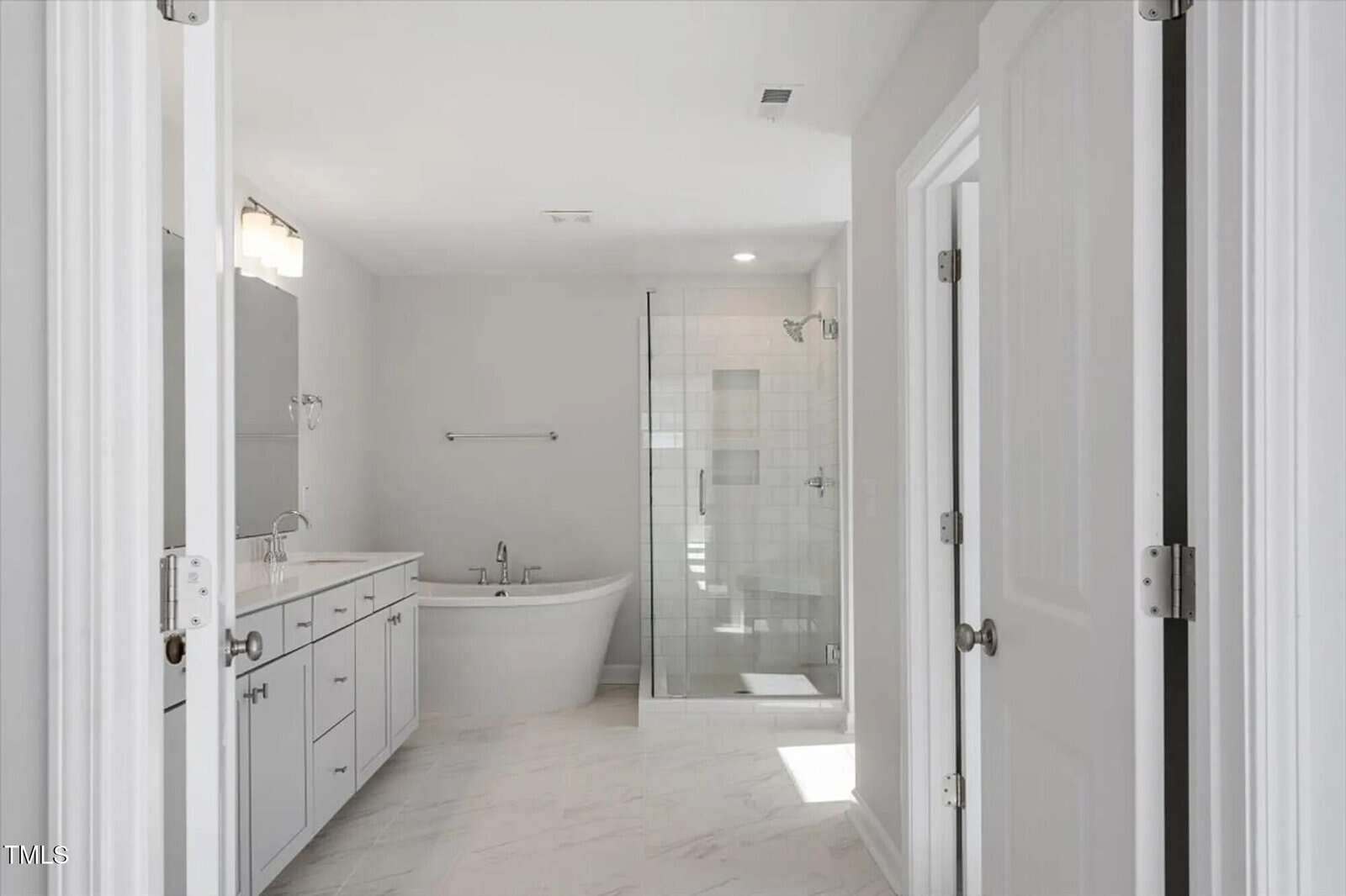 A modern bathroom with white walls, a double-sink vanity on the left, a freestanding bathtub in the center, and a glass-enclosed shower on the right. The floor is tiled in a light color.