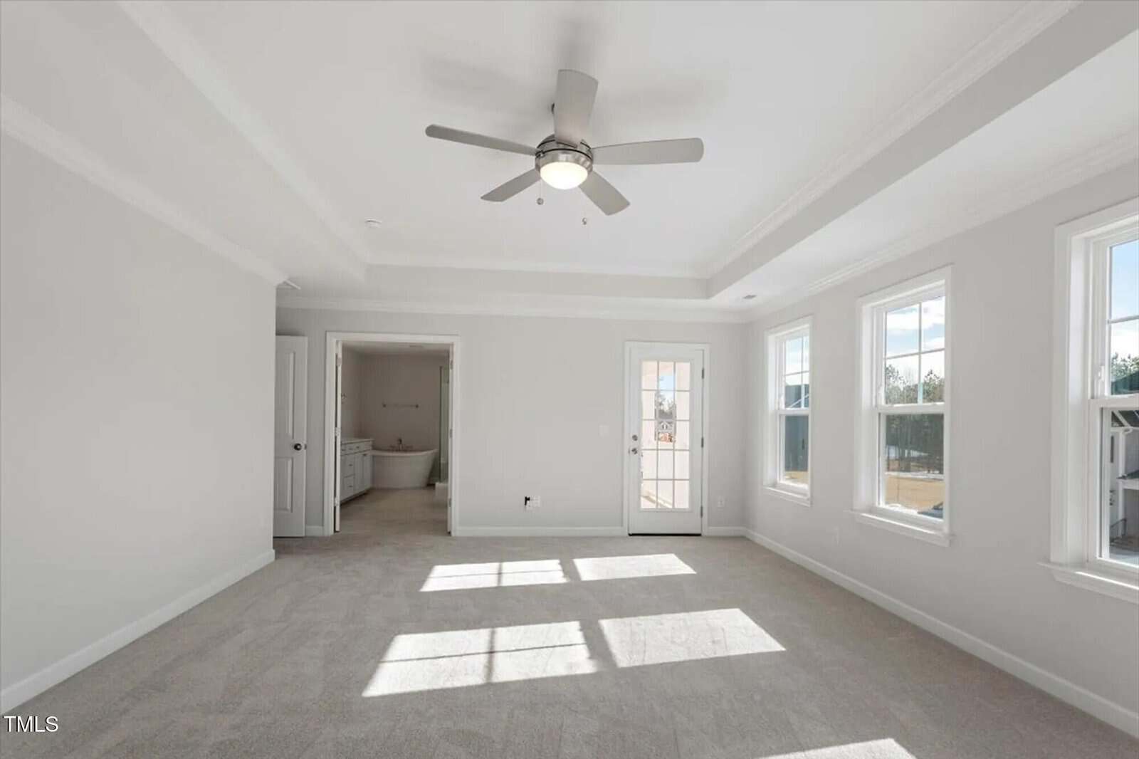 Bright, empty room with beige carpet, ceiling fan, and three large windows. An open door leads to a bathroom with a bathtub. Another door opens to the outside. Walls are painted light gray. Sunlight shines on the floor.