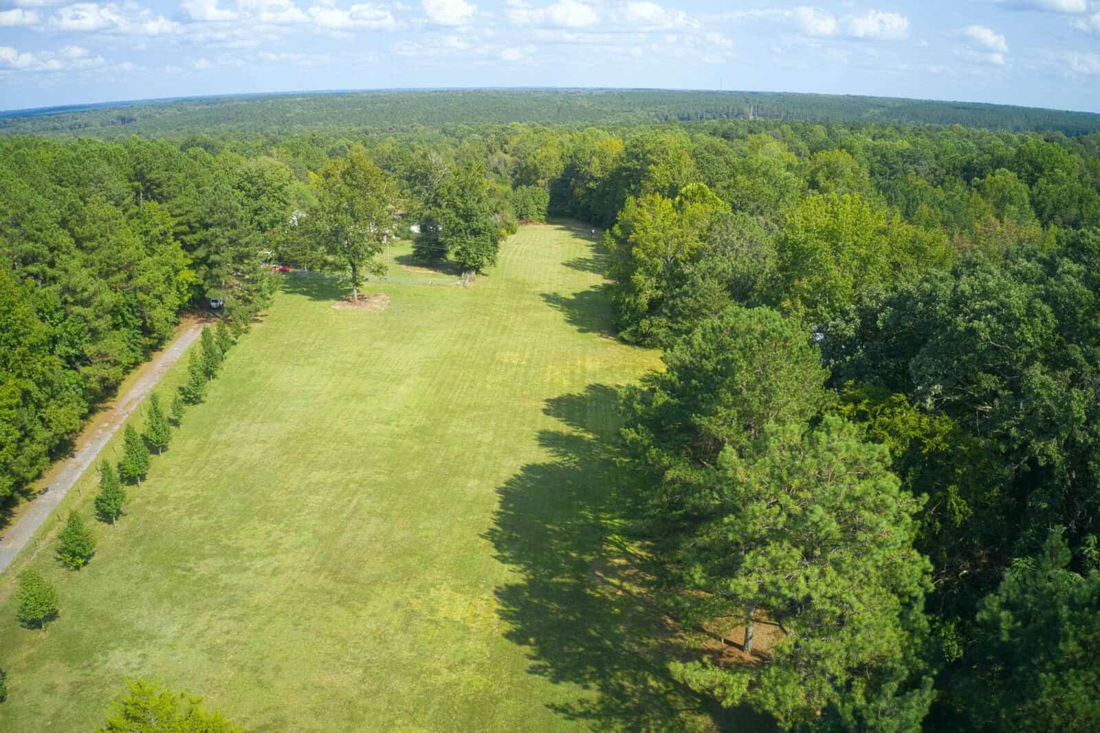 A large, grassy field bordered by tall trees on both sides, with a clear blue sky and scattered clouds overhead stretches peacefully near Franklinton. A gravel path runs along the left edge of the field at 878 Green Hill Road, leading into a wooded area.