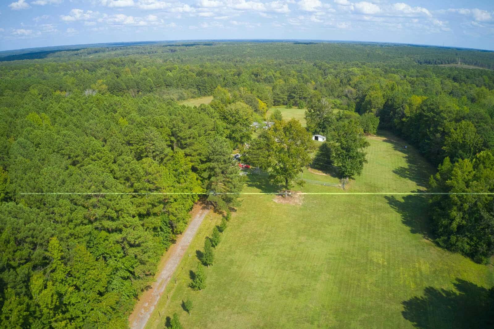 Aerial view of a large, grassy field bordered by dense forest, with a dirt road winding through the trees and a few scattered buildings visible in the distance under a partly cloudy sky.