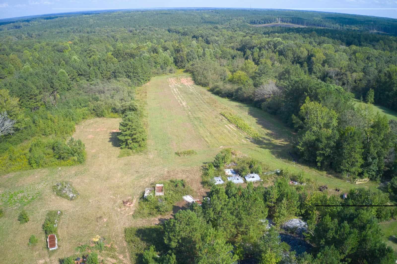 Aerial view of a large, grassy clearing surrounded by dense forest. Several small structures or objects are clustered near the bottom, with patches of trees and open fields visible throughout the landscape.